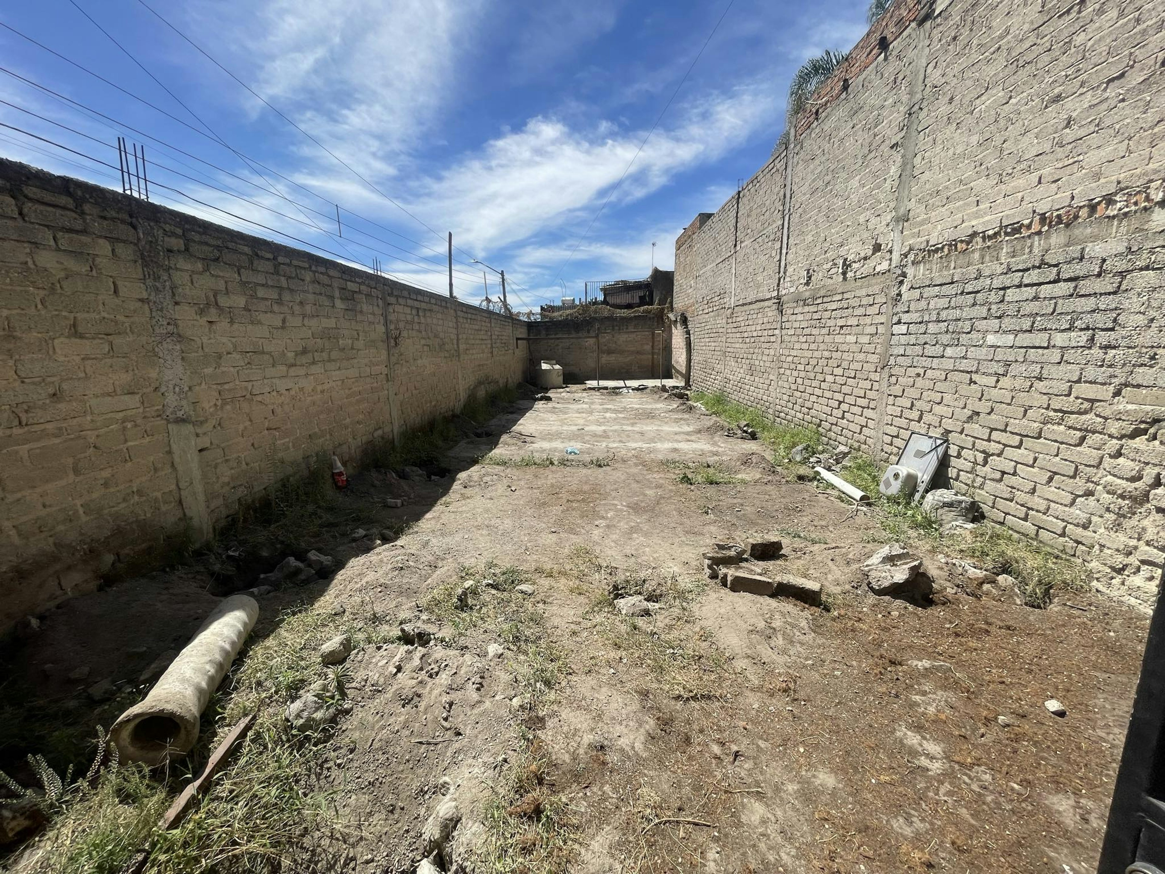 La imagen muestra un terreno baldío rodeado por muros de ladrillo altos. El suelo es mayormente de tierra, con algunos escombros y vegetación dispersa. Se observa un tubo de concreto en el suelo. Los muros tienen cables eléctricos por encima. El cielo es azul con algunas nubes. El lugar parece estar en una zona urbana en desarrollo o abandonada, posiblemente destinado para construcción futura.