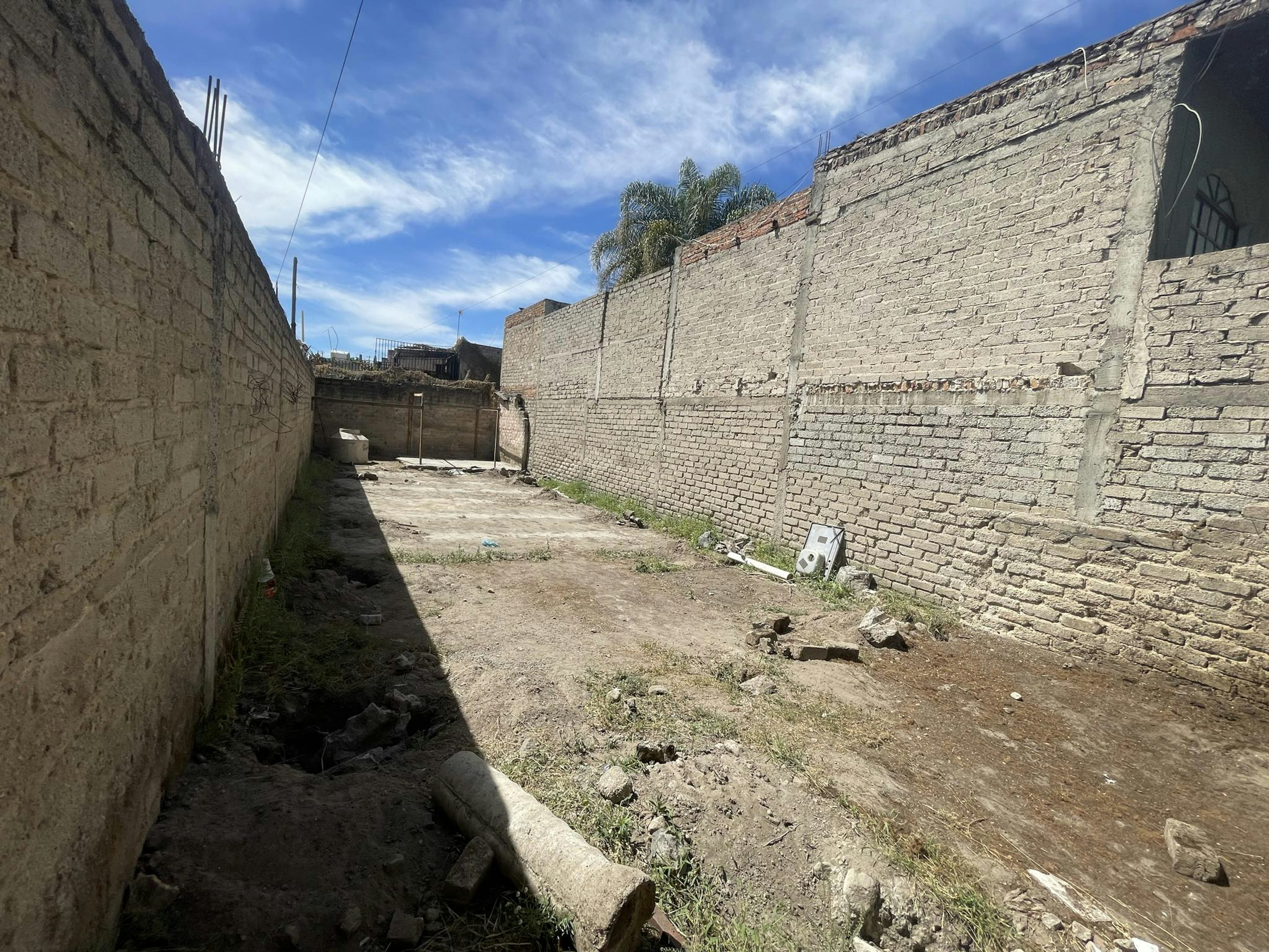 La imagen muestra un terreno baldío estrecho y alargado, rodeado por altos muros de ladrillo y concreto. El suelo es de tierra y está cubierto de escombros y vegetación dispersa. Se observa un cielo azul despejado y algunas estructuras en construcción al fondo. El espacio parece estar en una zona urbana en desarrollo, posiblemente destinado para futura construcción o ampliación de viviendas.