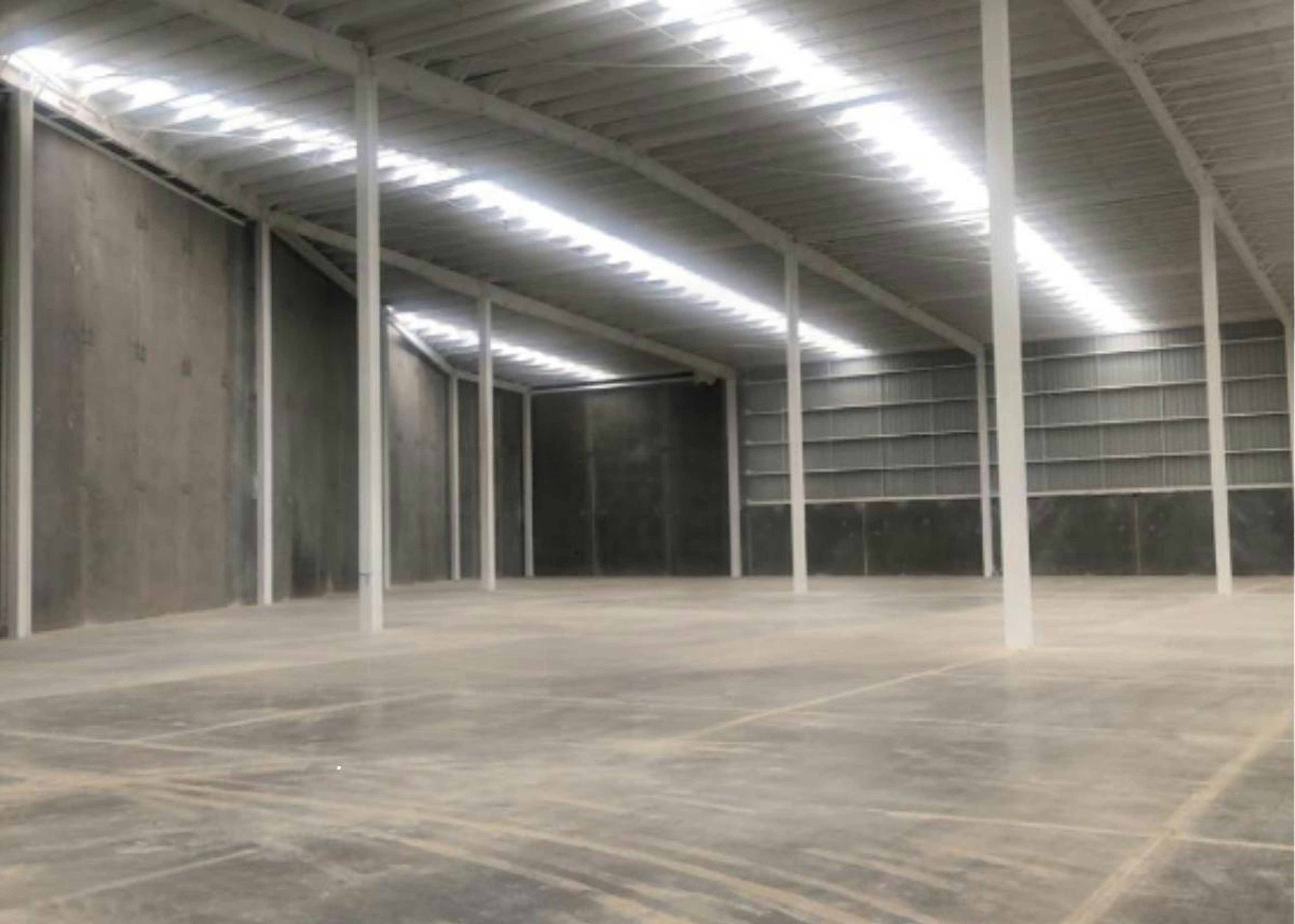 La imagen muestra un amplio espacio industrial con techo alto y estructura de vigas blancas. El piso es de concreto pulido y hay estanterías metálicas en las paredes. Se observa un logo translúcido superpuesto que sugiere Strategic Industry Logistics. El espacio parece ser un almacén o centro de distribución moderno, ideal para operaciones logísticas o industriales de gran escala.