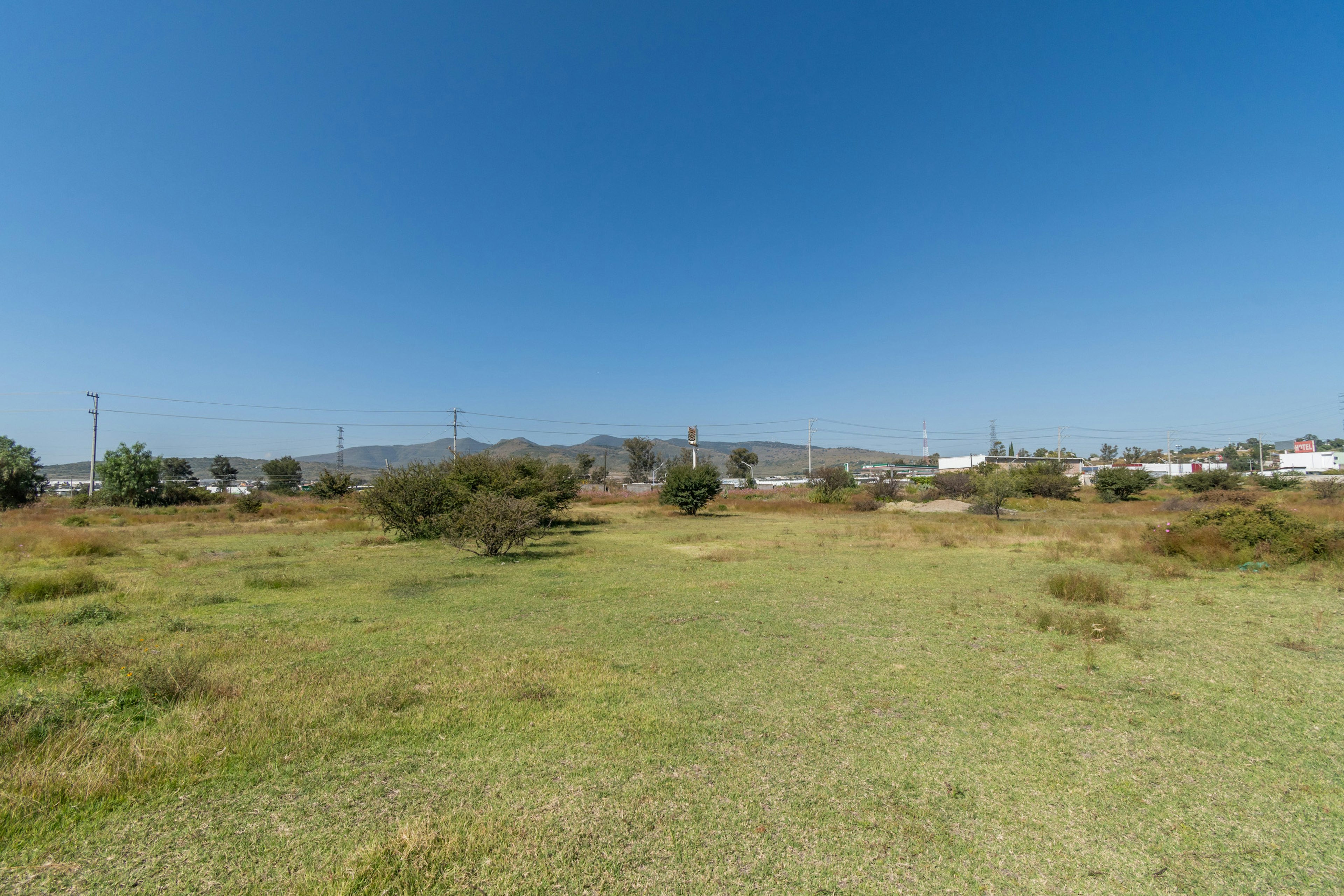 La imagen muestra un terreno amplio y llano con vegetación baja, ideal para desarrollo inmobiliario. Al fondo se observan montañas, líneas eléctricas y algunas construcciones, sugiriendo una zona periurbana en desarrollo. El cielo despejado y el paisaje abierto indican un clima favorable. Este tipo de terreno podría ser atractivo para proyectos residenciales o comerciales, aprovechando la vista y el espacio disponible.