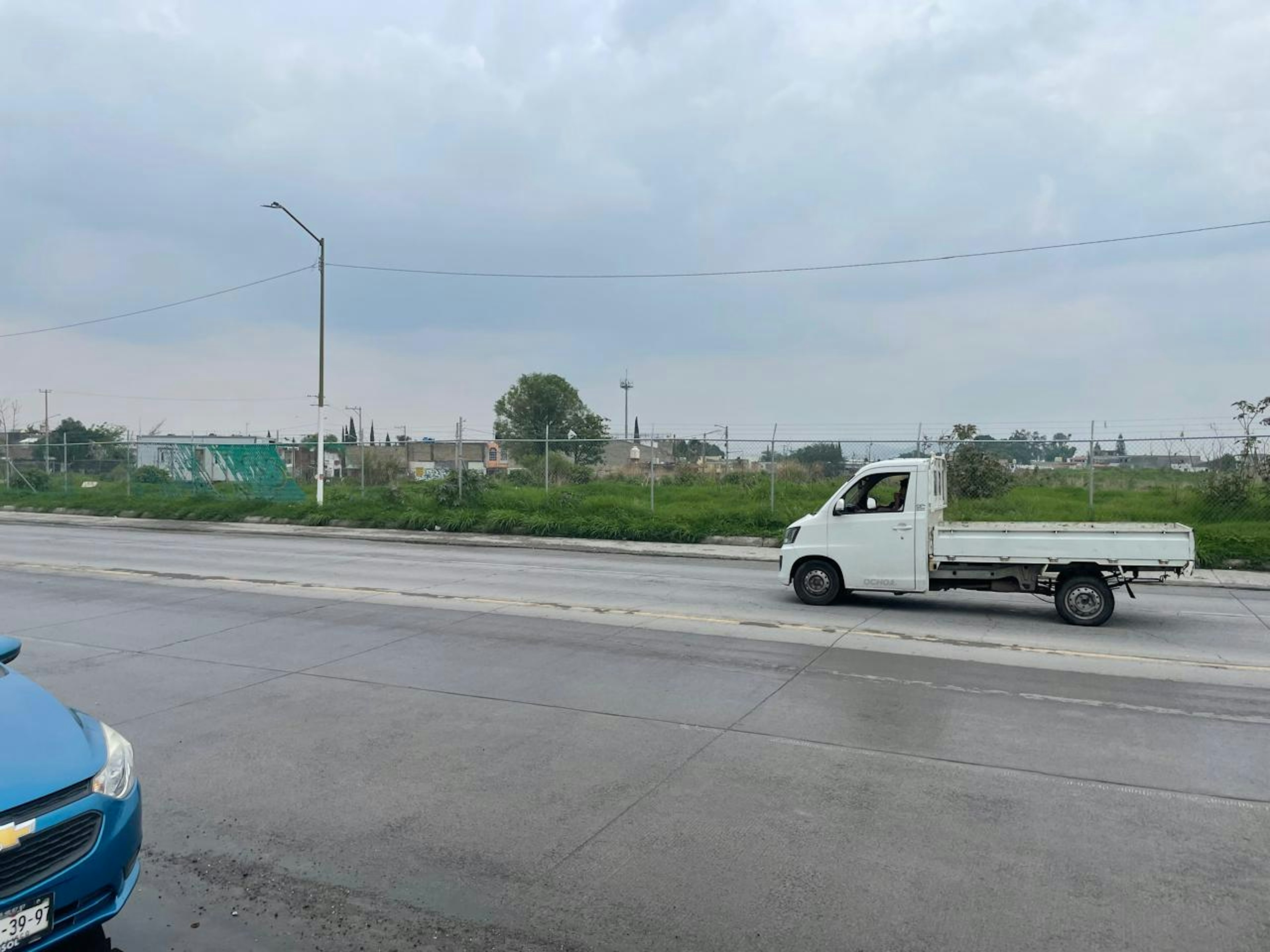 La imagen muestra una zona suburbana en desarrollo con un amplio terreno baldío al fondo. Se observa una calle pavimentada en primer plano, con postes de luz y cables eléctricos. Hay un vehículo utilitario blanco estacionado a un lado. El área parece estar en proceso de urbanización, con potencial para futuros desarrollos inmobiliarios o comerciales.