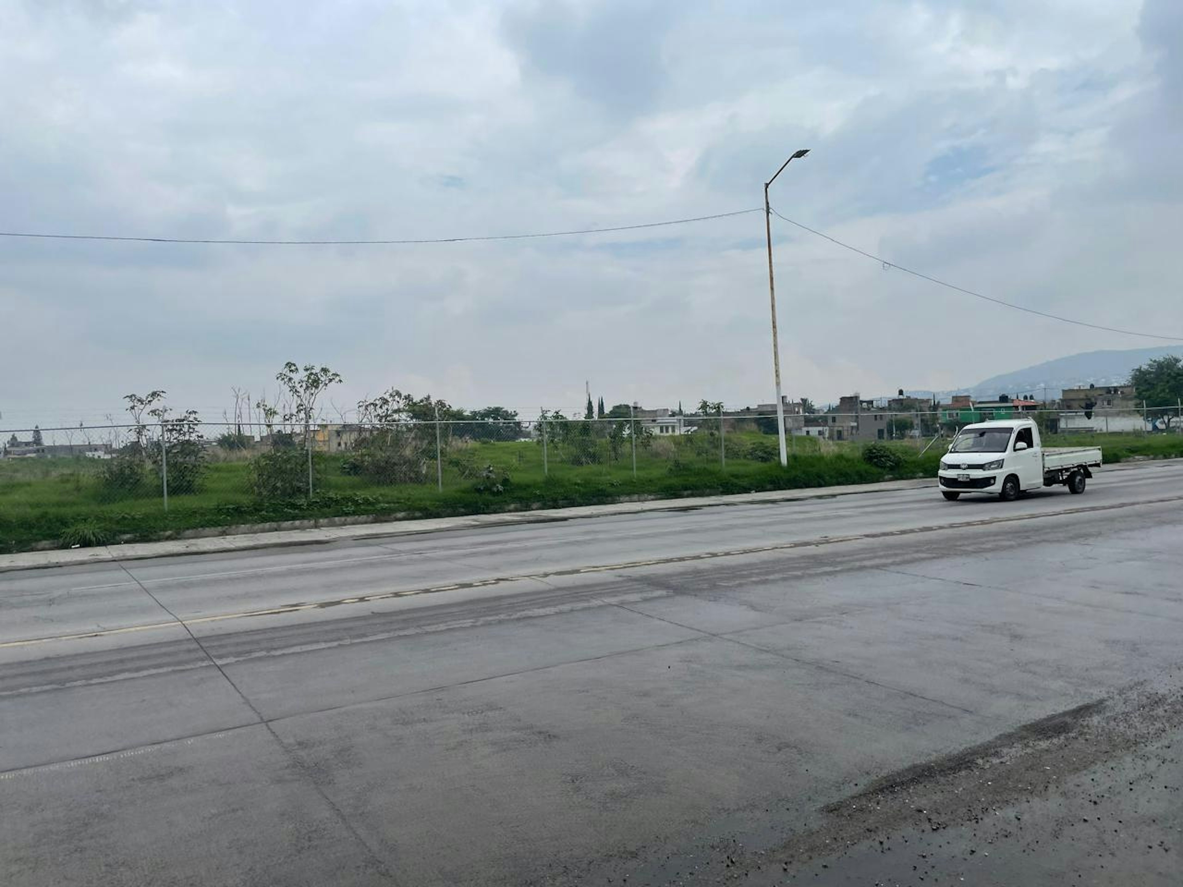 La imagen muestra una zona suburbana en desarrollo con una amplia carretera de concreto en primer plano. Al fondo se observa un área verde con vegetación y una cerca, seguida de edificaciones bajas y dispersas. Se aprecia un poste de luz y cables eléctricos. El cielo está nublado y se ve una pequeña camioneta blanca en la carretera. El terreno parece tener potencial para futuros desarrollos inmobiliarios.