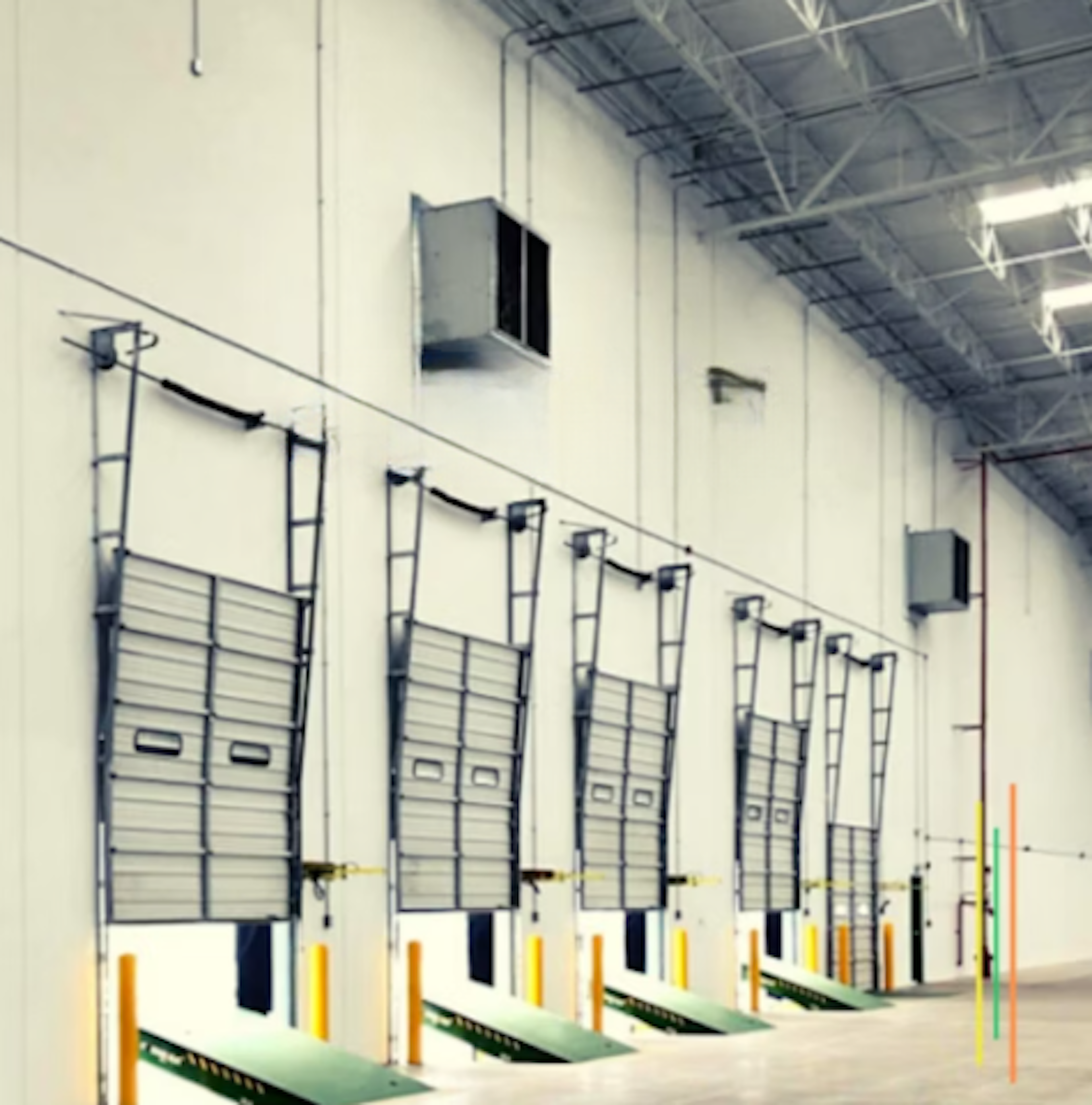 La imagen muestra una serie de puertas de carga industriales en un almacén o centro de distribución. Las puertas metálicas están alineadas a lo largo de una pared blanca, con sistemas de elevación visibles. Hay postes de seguridad amarillos y naranjas frente a cada puerta, así como rampas de acceso verdes. El techo alto con estructura expuesta y unidades de climatización sugiere un espacio logístico moderno y funcional.