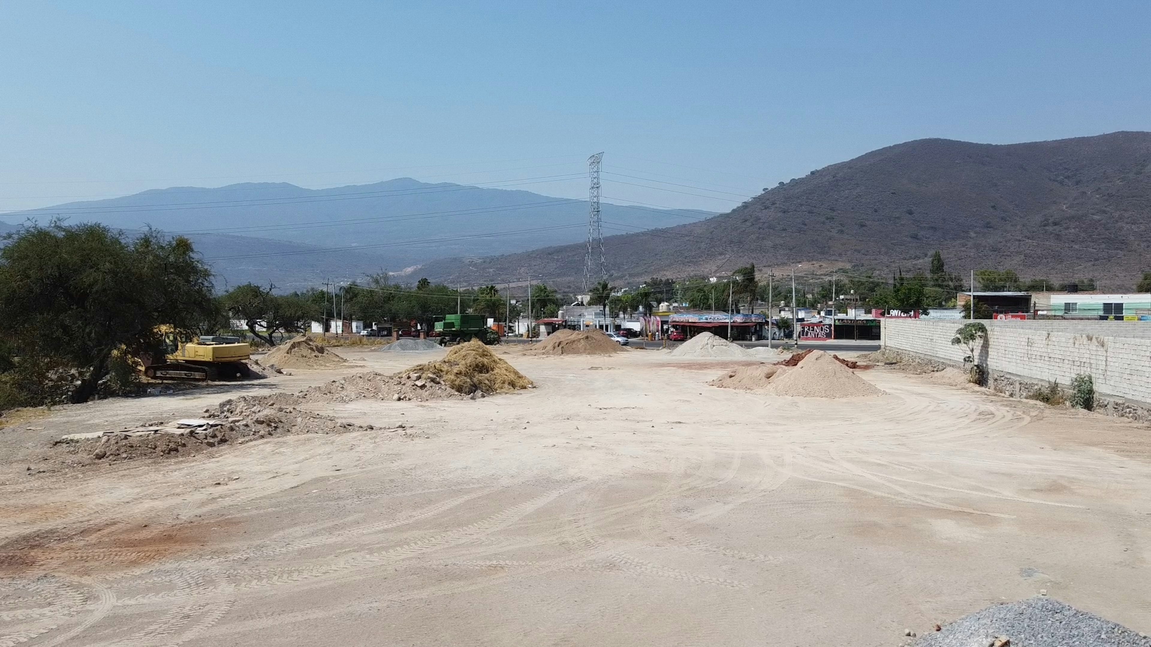 La imagen muestra un terreno en desarrollo en una zona semiárida. Se observa un área extensa de tierra despejada con montículos de arena y grava, sugiriendo preparación para construcción. Al fondo se ven edificaciones modestas y montañas, indicando una ubicación periurbana. Hay infraestructura eléctrica visible. El paisaje árido y la presencia de maquinaria pesada sugieren un proyecto inmobiliario en sus etapas iniciales.