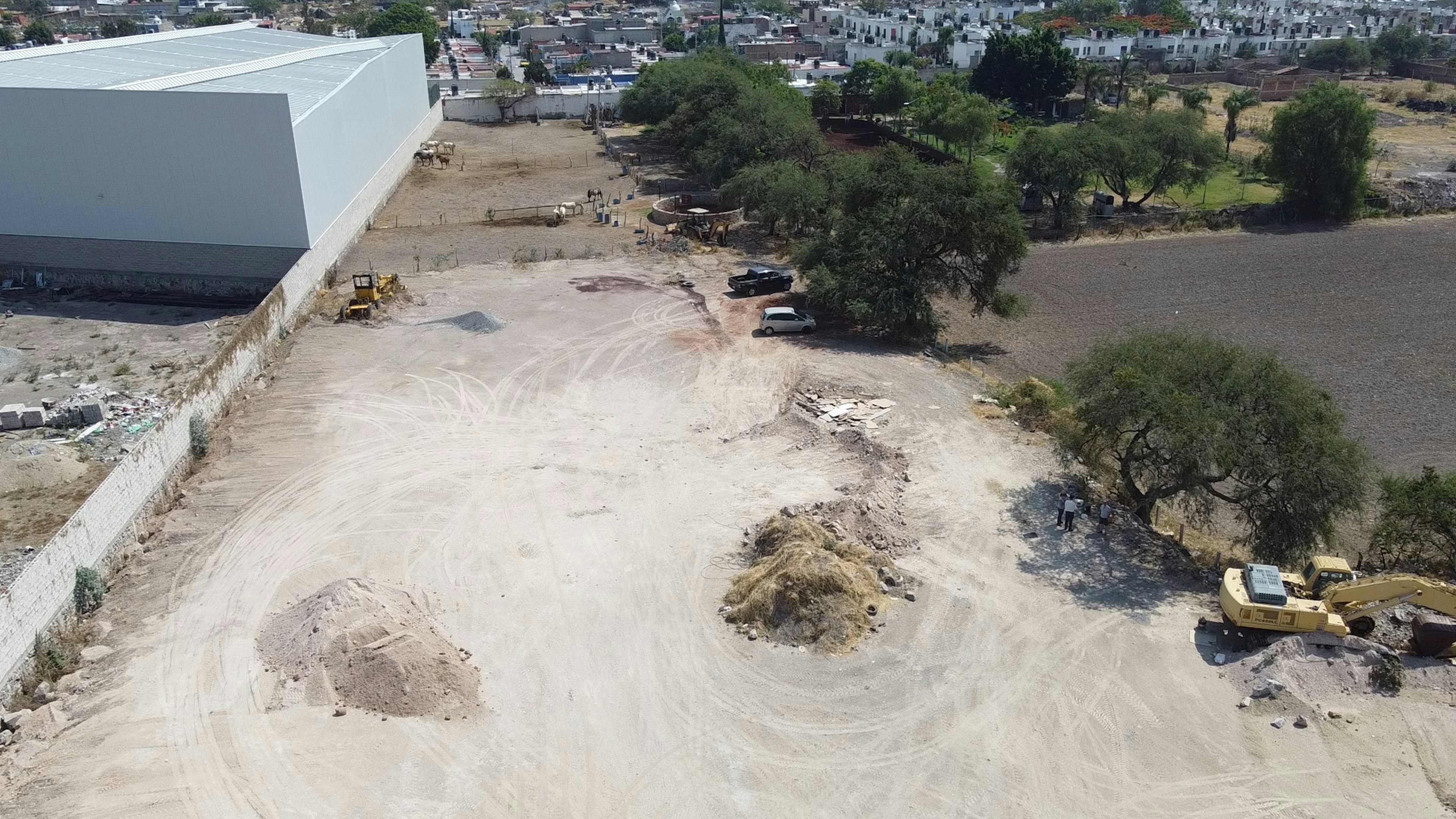 La imagen muestra un área en desarrollo con un gran edificio blanco a la izquierda. En primer plano hay un terreno despejado con marcas de vehículos, montículos de tierra y equipo de construcción. Se observan algunos árboles dispersos y vehículos estacionados. Al fondo se ve una zona residencial con casas blancas y vegetación. El área parece estar en proceso de urbanización o construcción.