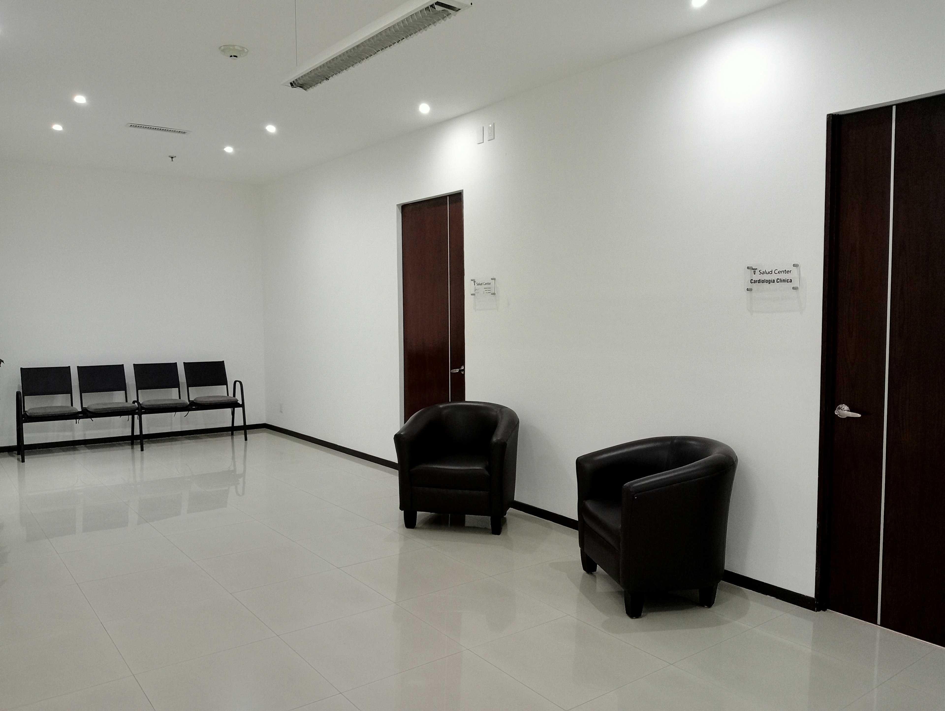 Espacio de oficina moderno y minimalista con iluminación empotrada, suelo de baldosas brillantes, dos sillones de cuero y una zona de espera con asientos. Ideal para consultorios o empresas.