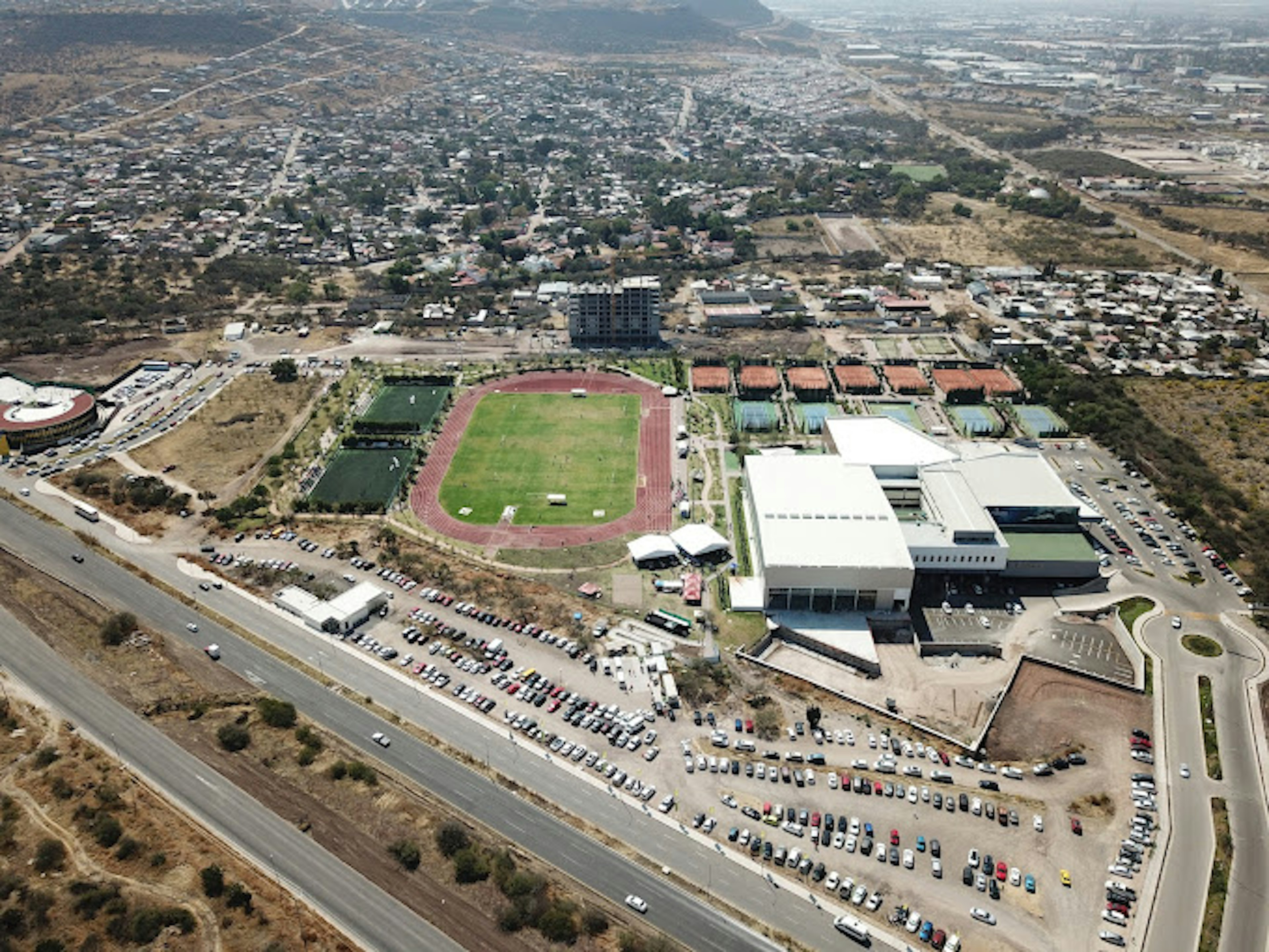 La imagen muestra una vista aérea de un complejo deportivo con un estadio de atletismo, campos de fútbol y canchas de tenis. Alrededor hay un amplio estacionamiento lleno de vehículos. El complejo está rodeado por una zona urbana en desarrollo con edificios residenciales y comerciales. Se observa una carretera principal que pasa junto al complejo. El área circundante parece semiárida, con terrenos sin desarrollar en las afueras de la ciudad.