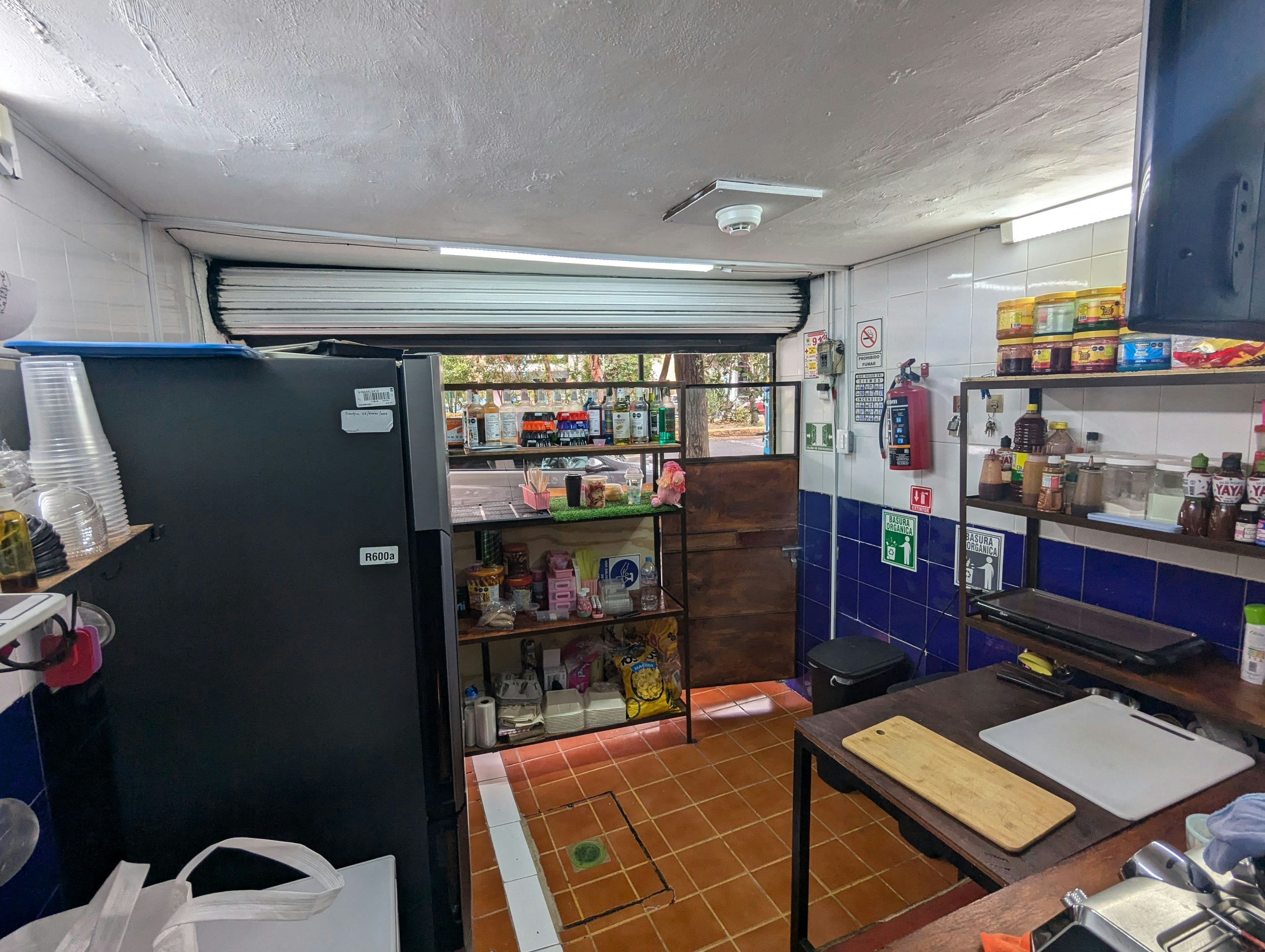 La imagen muestra el interior de un pequeño negocio o tienda, posiblemente un kiosco o minimercado. Se observa un refrigerador, estantes con productos variados, una pequeña cocina y un mostrador. El espacio es compacto, con paredes de azulejos blancos y azules, y un suelo de baldosas naranjas. Hay signos de seguridad y un extintor visible, indicando que es un local comercial. La puerta enrollable sugiere que es un negocio que se abre al público.