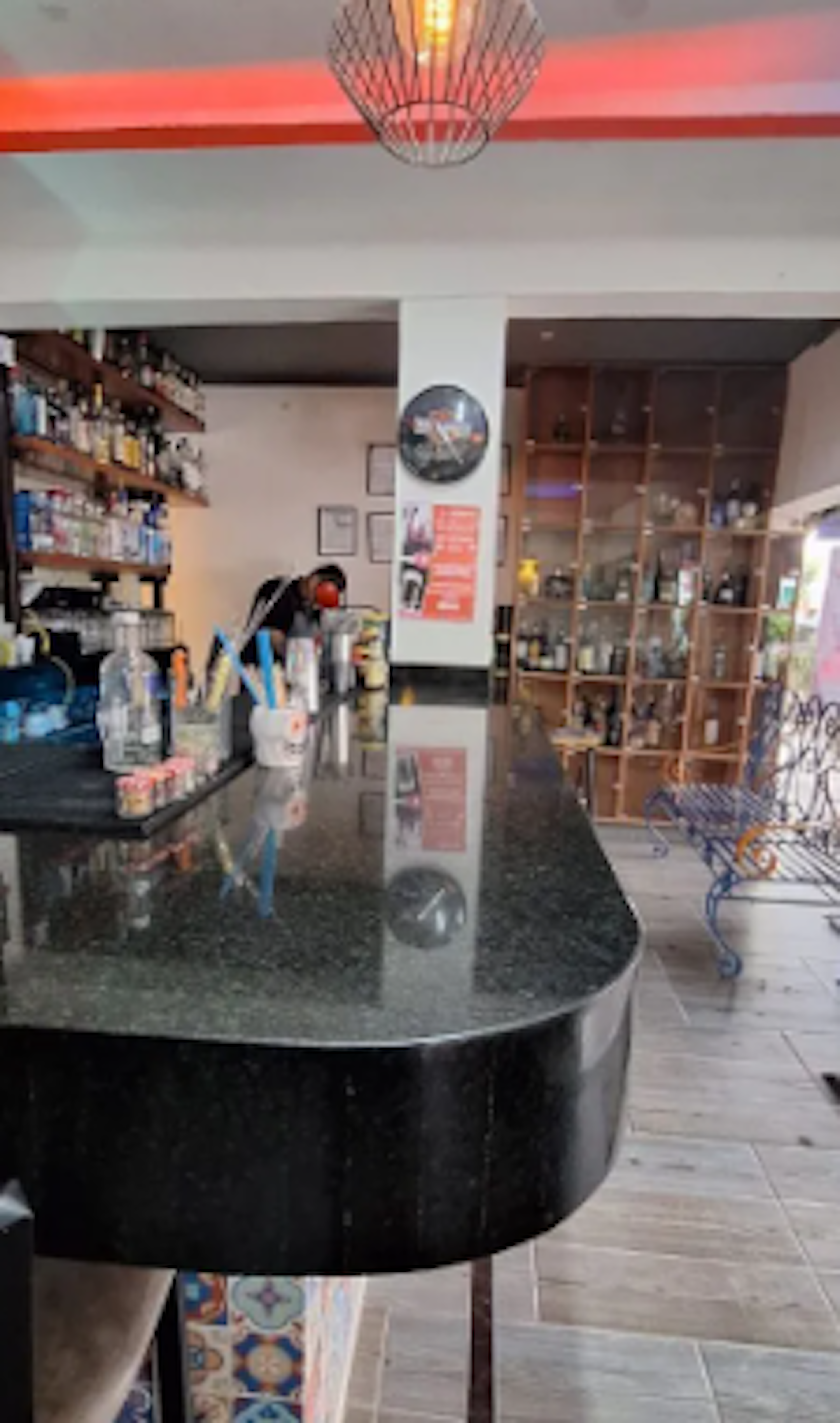 La imagen muestra el interior de un bar o cafetería con un mostrador de granito negro en primer plano. Detrás hay estanterías llenas de botellas de licor y un área de almacenamiento. El techo tiene una luz roja y una lámpara colgante decorativa. El espacio parece acogedor y bien equipado para servir bebidas, con un ambiente moderno y elegante.