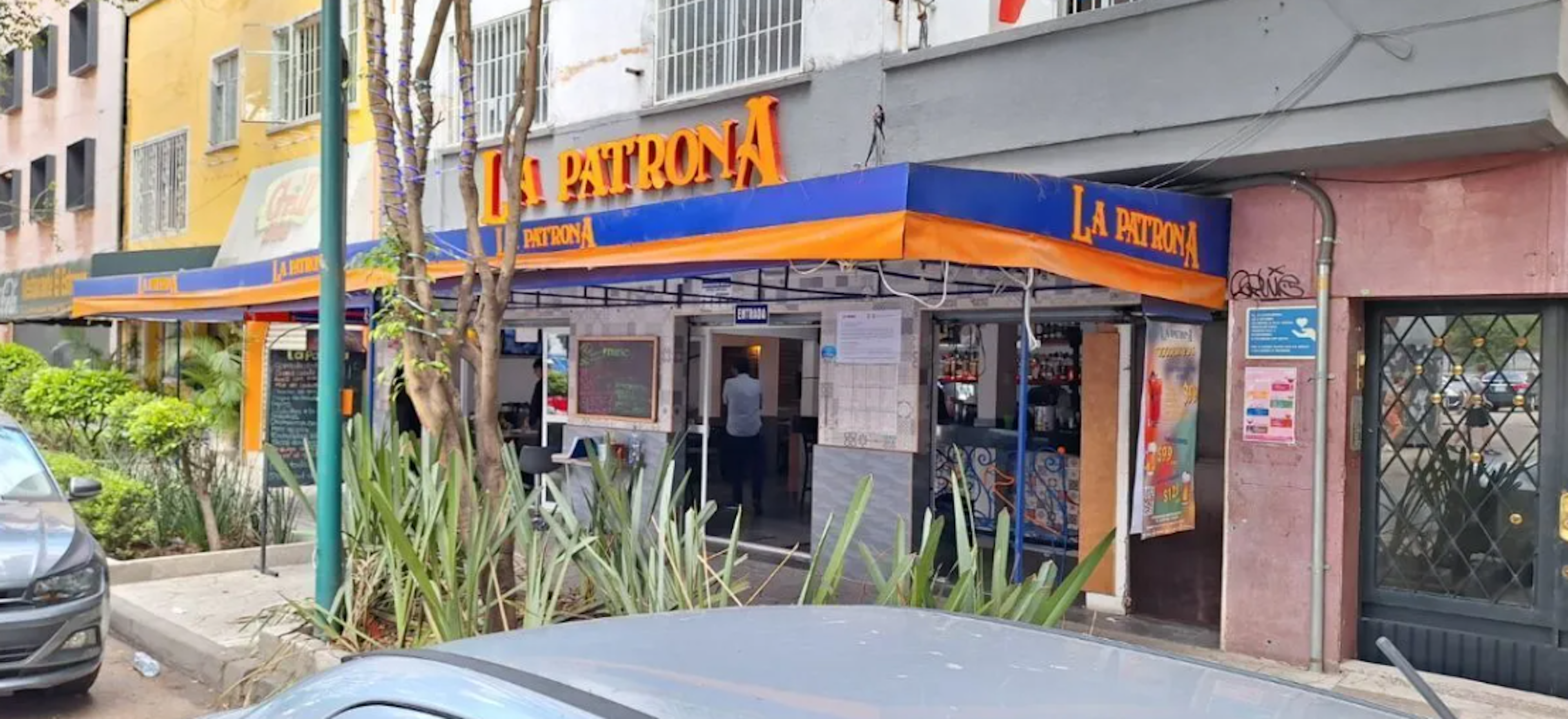 La imagen muestra la fachada de un restaurante llamado La Patrona en una zona urbana. El establecimiento tiene un toldo azul y naranja distintivo. Se encuentra en la planta baja de un edificio residencial, con otros negocios adyacentes. La calle tiene árboles y plantas, lo que sugiere un área comercial mezclada con viviendas en un barrio potencialmente atractivo para inversiones inmobiliarias.