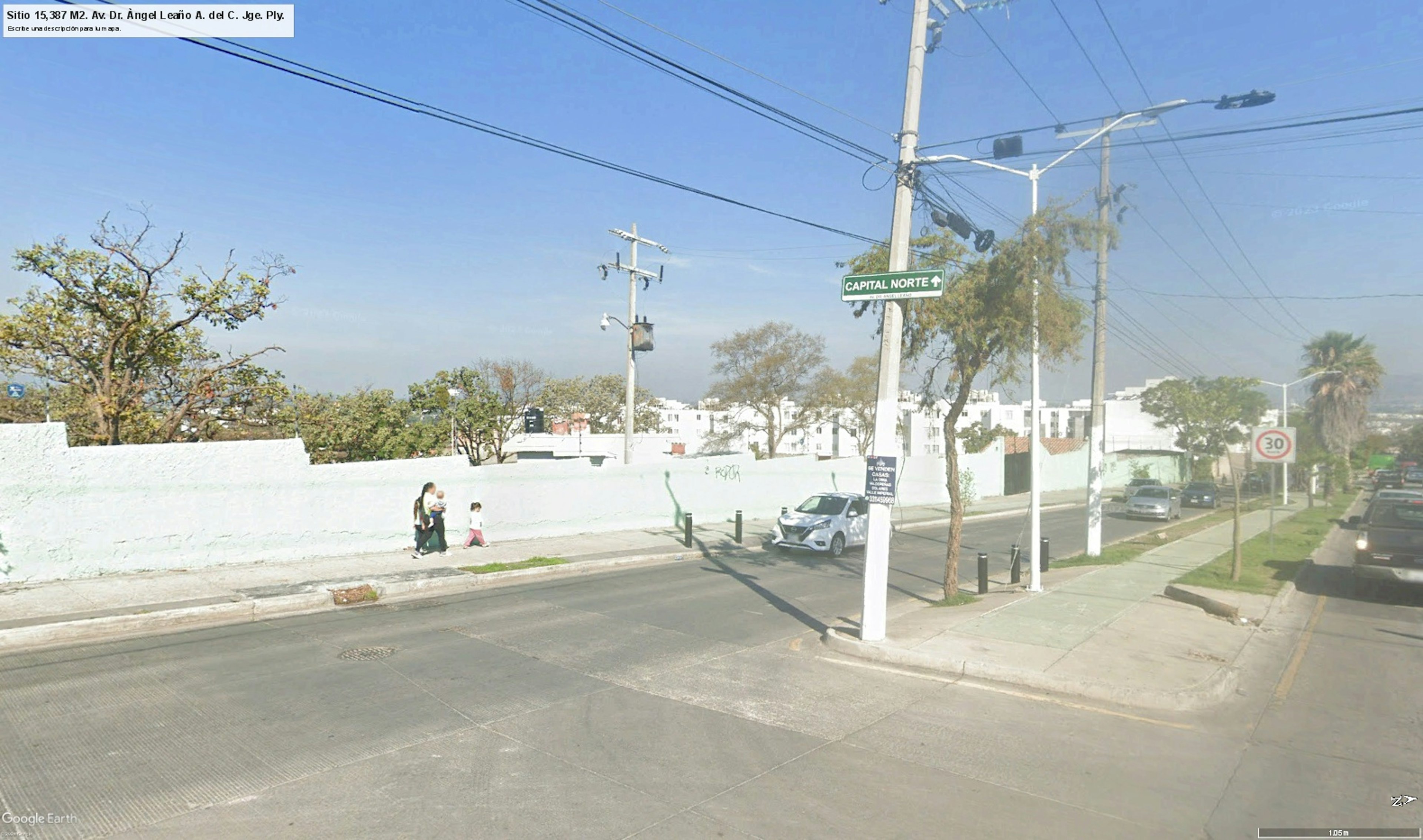 La imagen muestra una calle residencial en una zona urbana, posiblemente en México. Se observa una pared blanca larga que podría ser parte de un complejo residencial o condominio. Hay postes de electricidad, árboles y algunas personas caminando. La señal Capital Norte sugiere que es un área desarrollada. El entorno parece tranquilo y ordenado, típico de un barrio residencial de clase media.