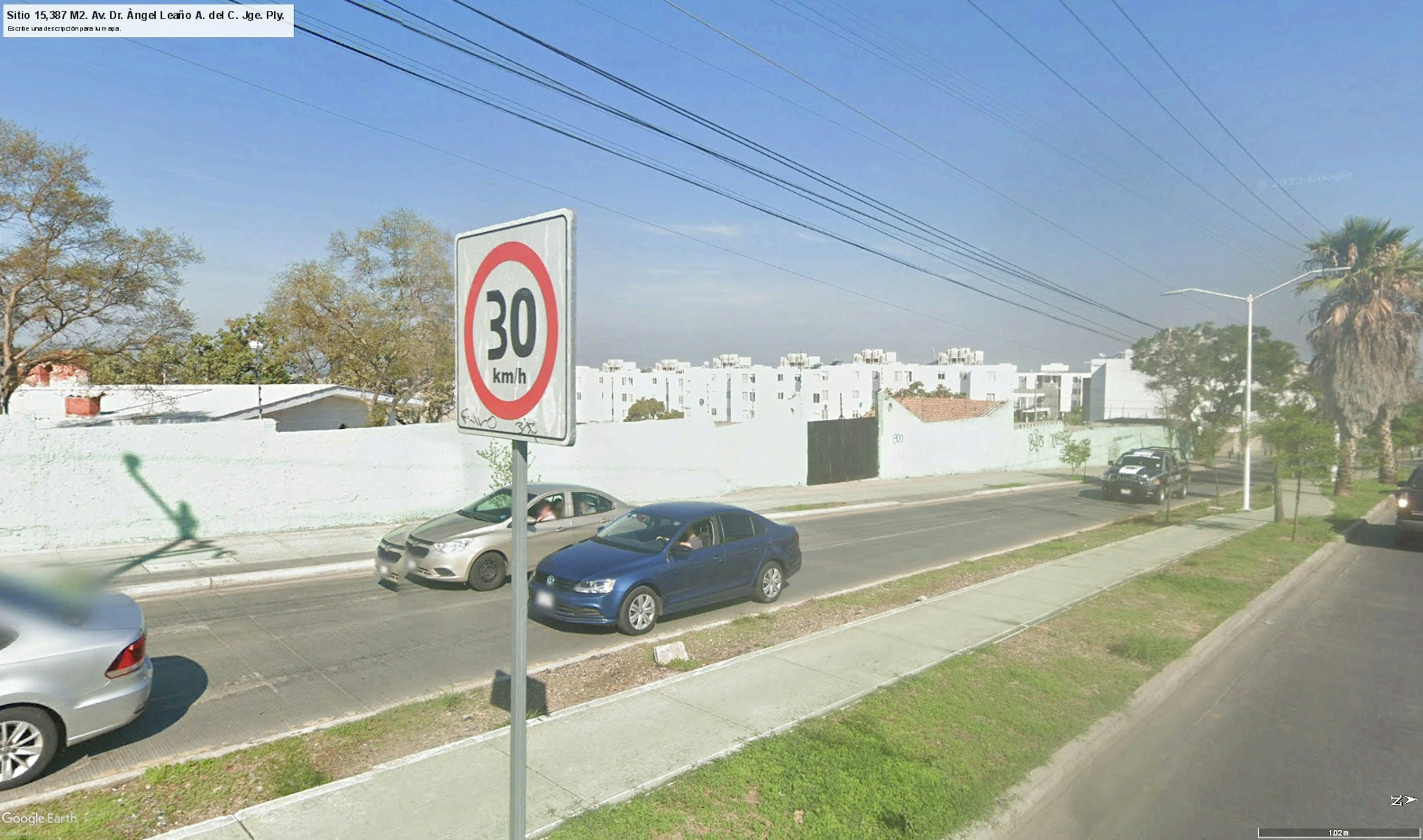 La imagen muestra una calle urbana con un límite de velocidad de 30 km/h. Se observan varios automóviles estacionados y en movimiento. Al fondo se ven edificios de apartamentos blancos, típicos de un desarrollo inmobiliario residencial. La escena incluye elementos urbanos como cables eléctricos, árboles y aceras, sugiriendo una zona residencial en desarrollo con infraestructura moderna.