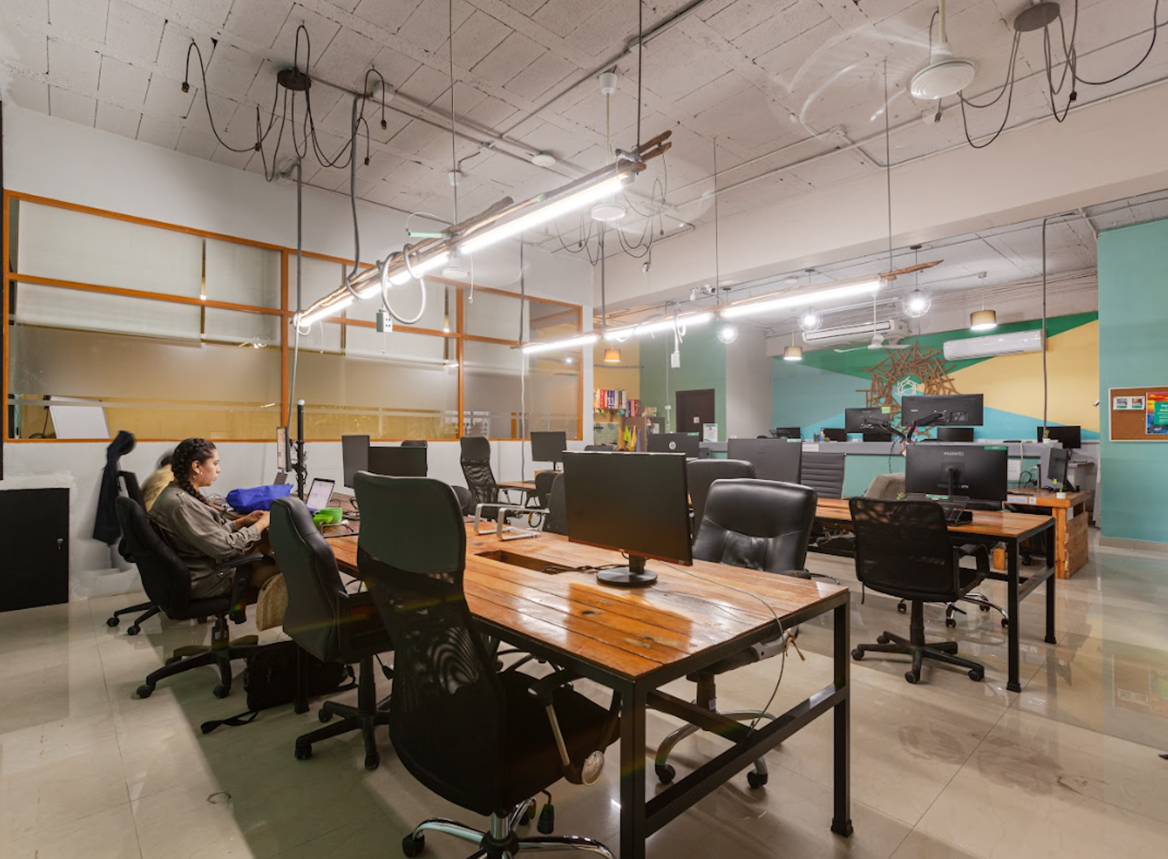 Espacio de oficina moderno y abierto con múltiples estaciones de trabajo y sillas ergonómicas. Iluminación colgante y decoración minimalista. Una persona está sentada trabajando en una computadora.