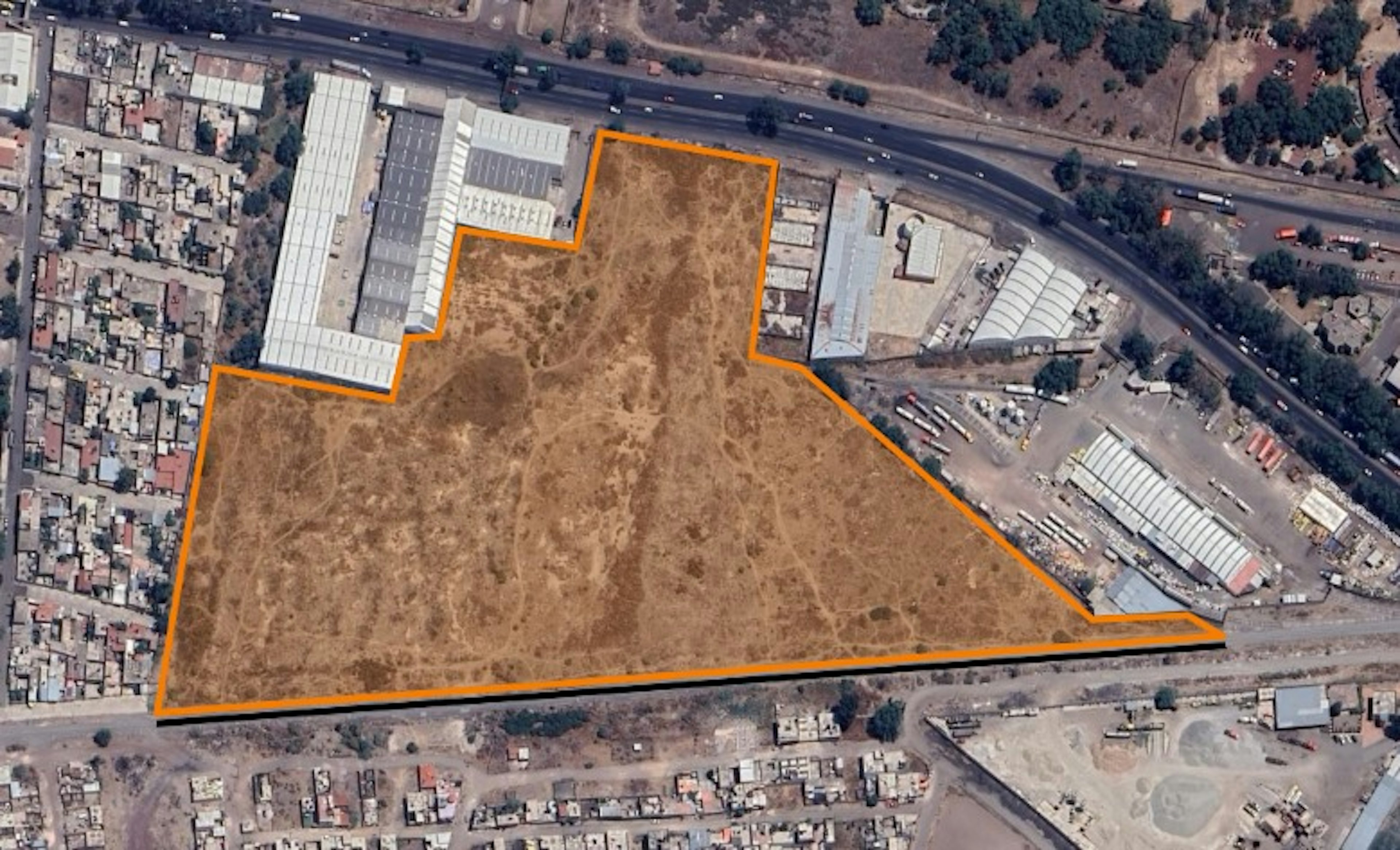 La imagen muestra una vista aérea de un terreno baldío de gran tamaño, delimitado por una línea naranja. El terreno está rodeado por zonas urbanizadas, incluyendo edificios industriales, almacenes y áreas residenciales. Su ubicación estratégica entre zonas desarrolladas sugiere un alto potencial para desarrollo inmobiliario, ya sea comercial, industrial o mixto.