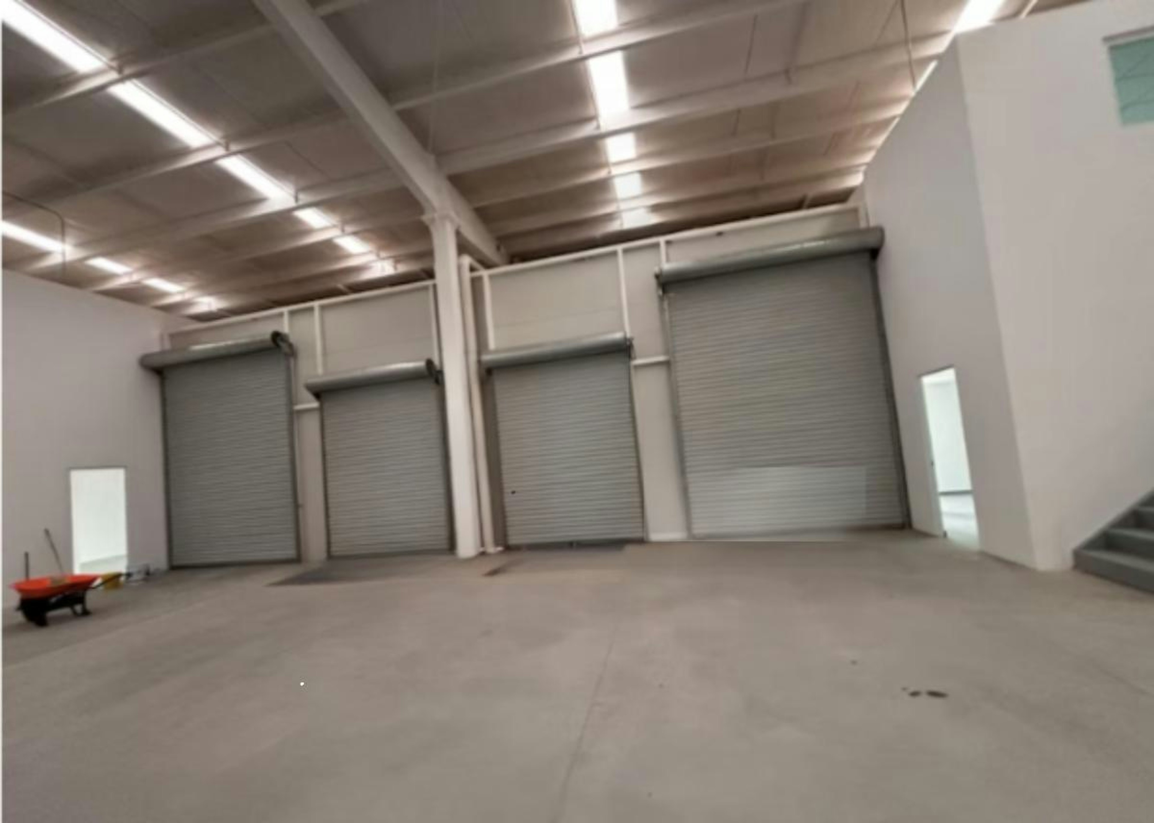 La imagen muestra un espacio industrial amplio y vacío con tres puertas enrollables metálicas cerradas. El techo es alto con iluminación fluorescente. El piso es de concreto liso. Se observa una carretilla roja en primer plano. Las paredes son blancas y hay algunas aberturas pequeñas. La imagen tiene una marca de agua que dice Strategic Industry Logistic Industry, sugiriendo que es un espacio para almacenamiento o distribución logística.