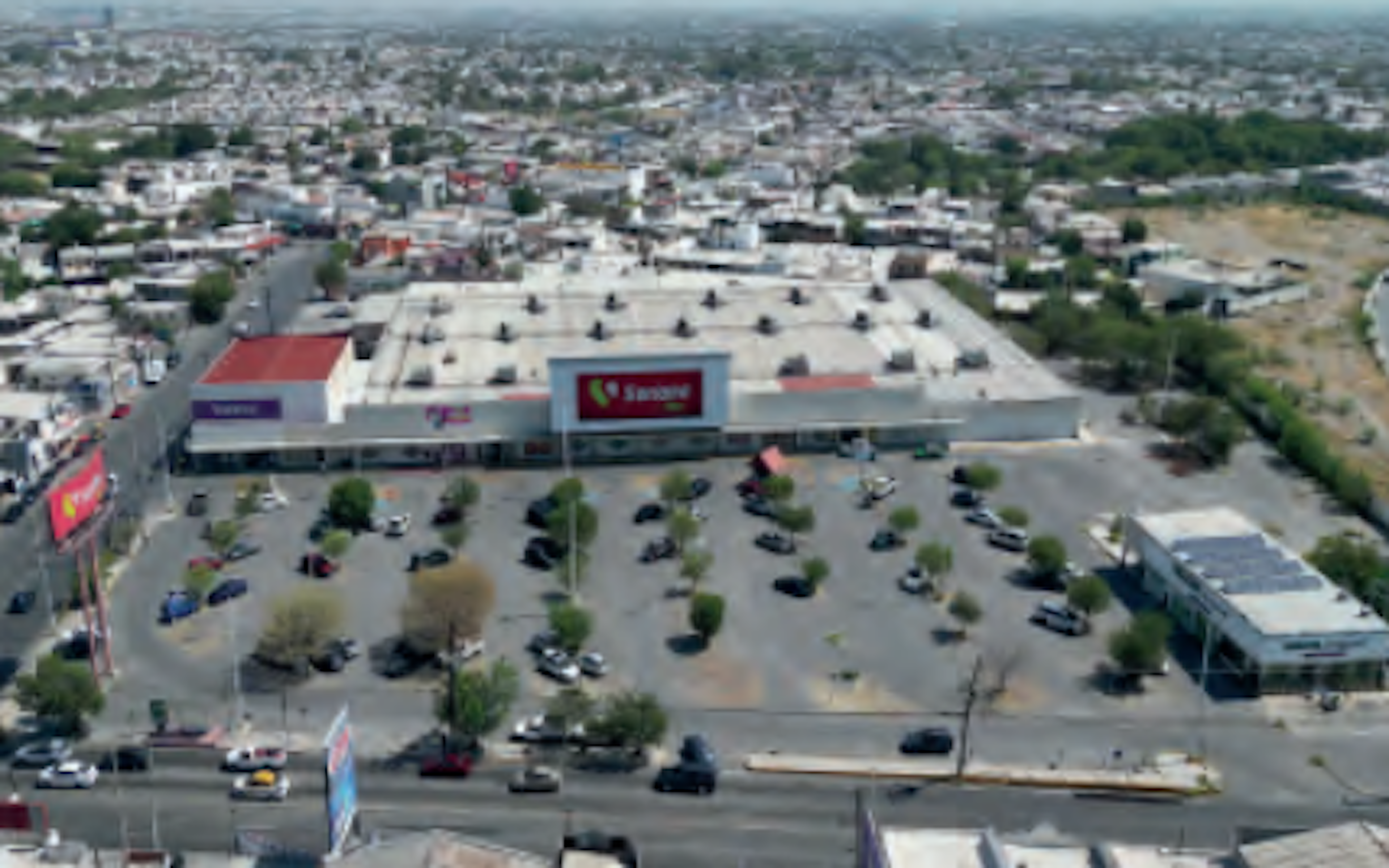 La imagen muestra una vista aérea de un gran centro comercial o supermercado, posiblemente una tienda Soriana, en medio de una zona urbana densamente poblada. El edificio blanco y rojo domina la escena, rodeado por un amplio estacionamiento con numerosos vehículos. El área circundante está llena de casas y edificios, lo que sugiere una ubicación céntrica en una ciudad mexicana. Este tipo de propiedad comercial es un activo inmobiliario significativo en áreas urbanas.