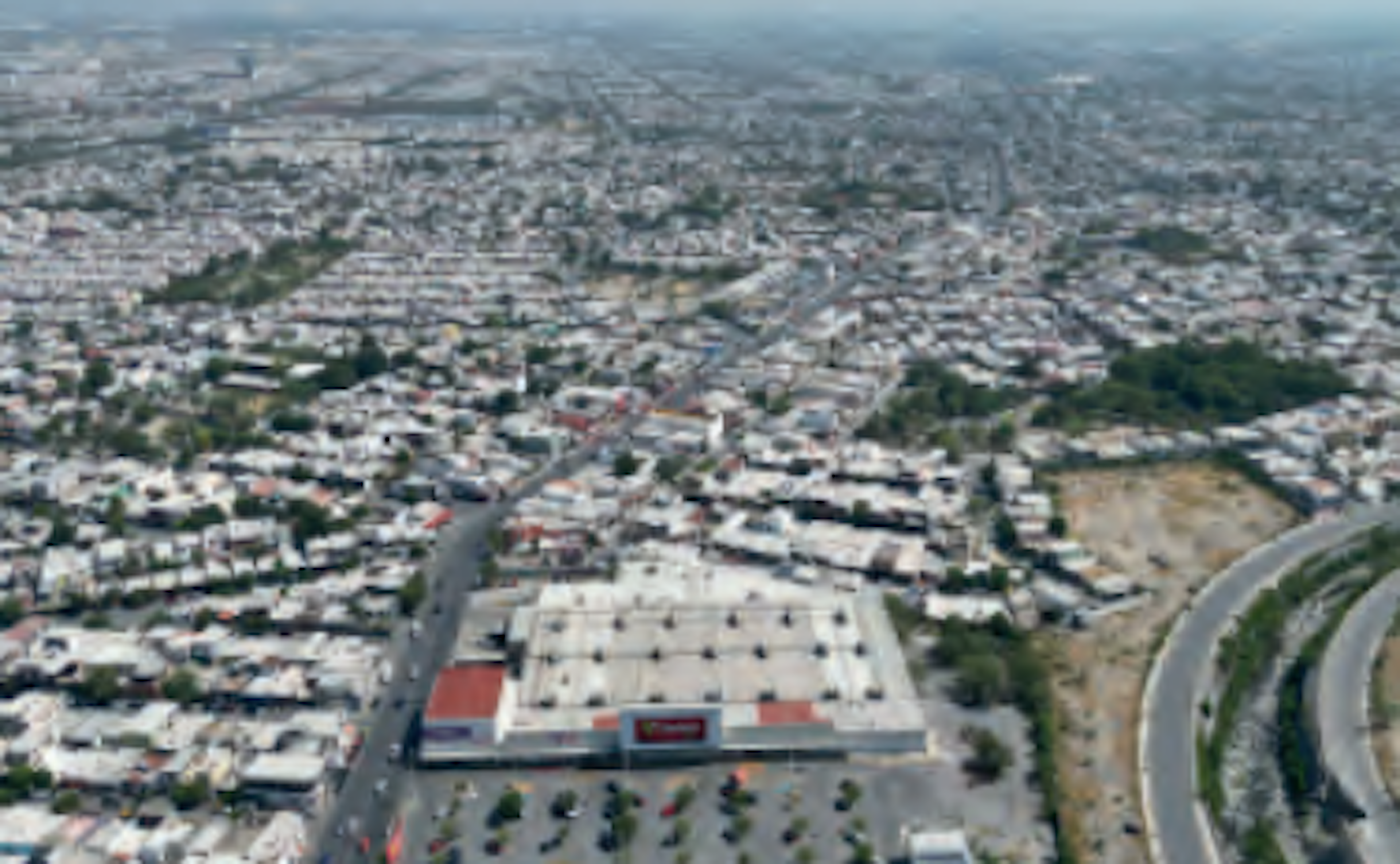 La imagen muestra una vista aérea de una extensa zona urbana densamente poblada. En el centro se aprecia un gran edificio comercial, posiblemente un centro comercial, con un amplio estacionamiento. La urbanización se extiende hasta el horizonte, con numerosas casas y edificios. Se observan varias vías de comunicación, incluyendo una carretera curva en primer plano. El paisaje es típico de una ciudad en desarrollo con una mezcla de áreas residenciales y comerciales.