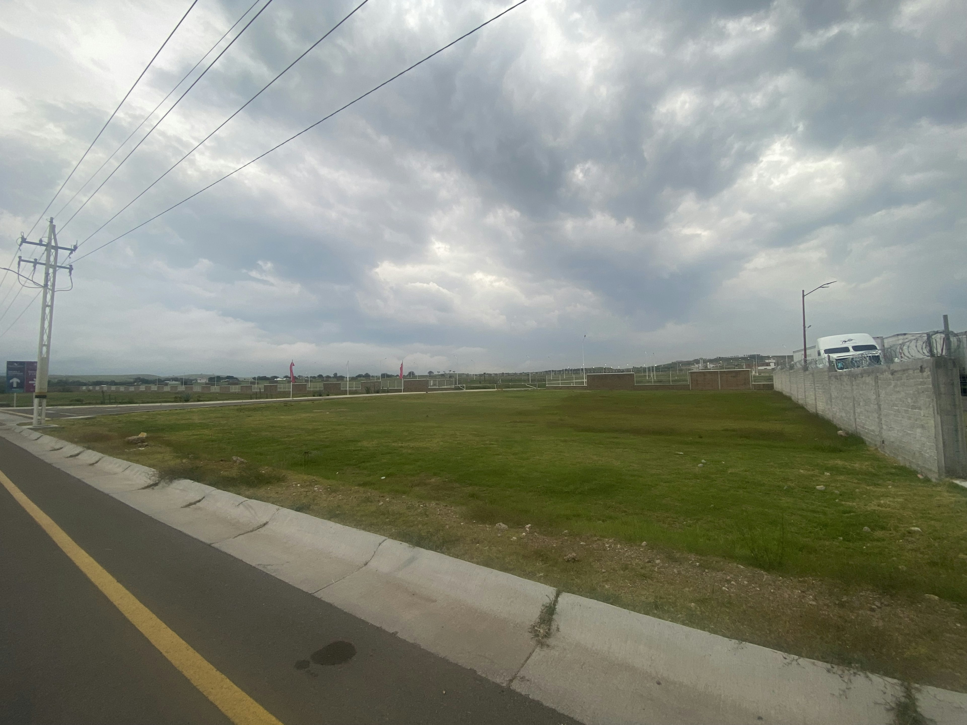 Esta imagen muestra un terreno vacío en una zona en desarrollo, con potencial para construcción inmobiliaria. Se observa césped verde, una carretera adyacente y cielo nublado.