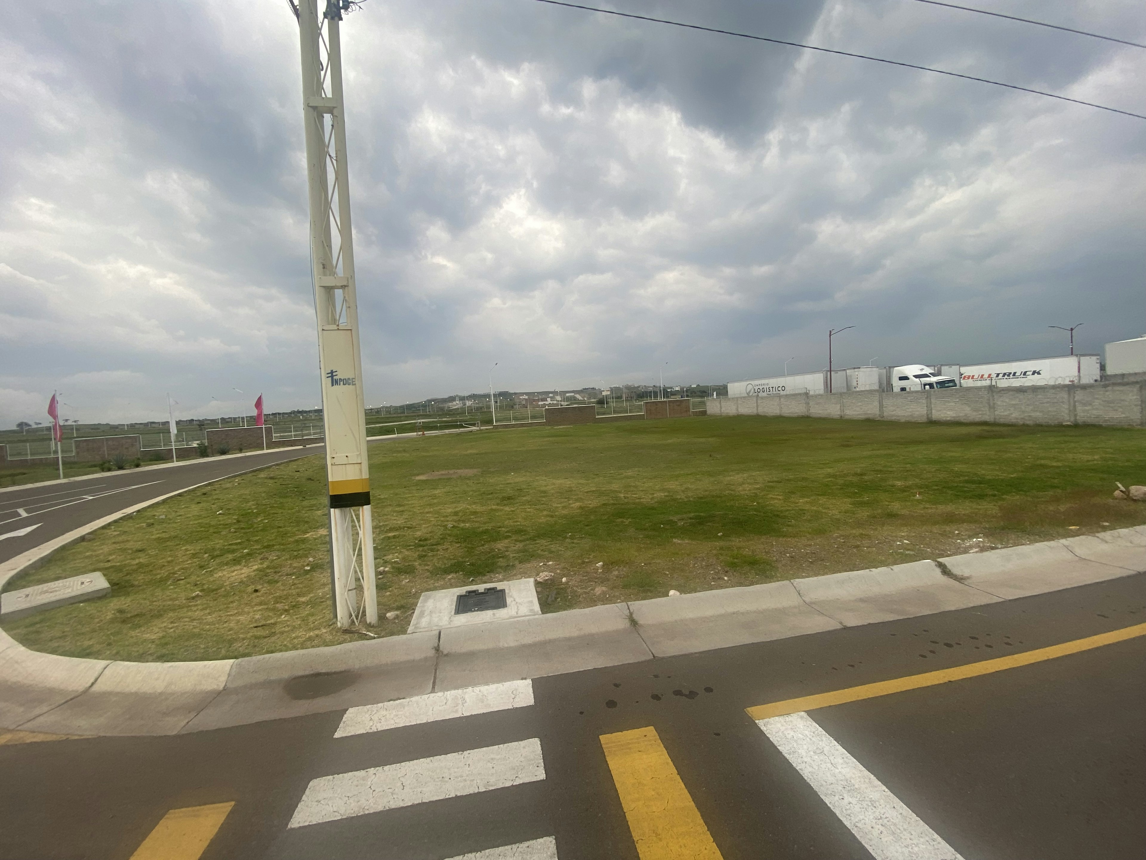 La imagen muestra un terreno vacío en una zona que parece estar en desarrollo, con infraestructura vial en primer plano y edificaciones industriales al fondo. El cielo está nublado, lo que sugiere un clima variable. Este espacio podría ser adecuado para futuros proyectos inmobiliarios, ya sea residenciales o comerciales, dada su aparente proximidad a vías de acceso y servicios existentes.