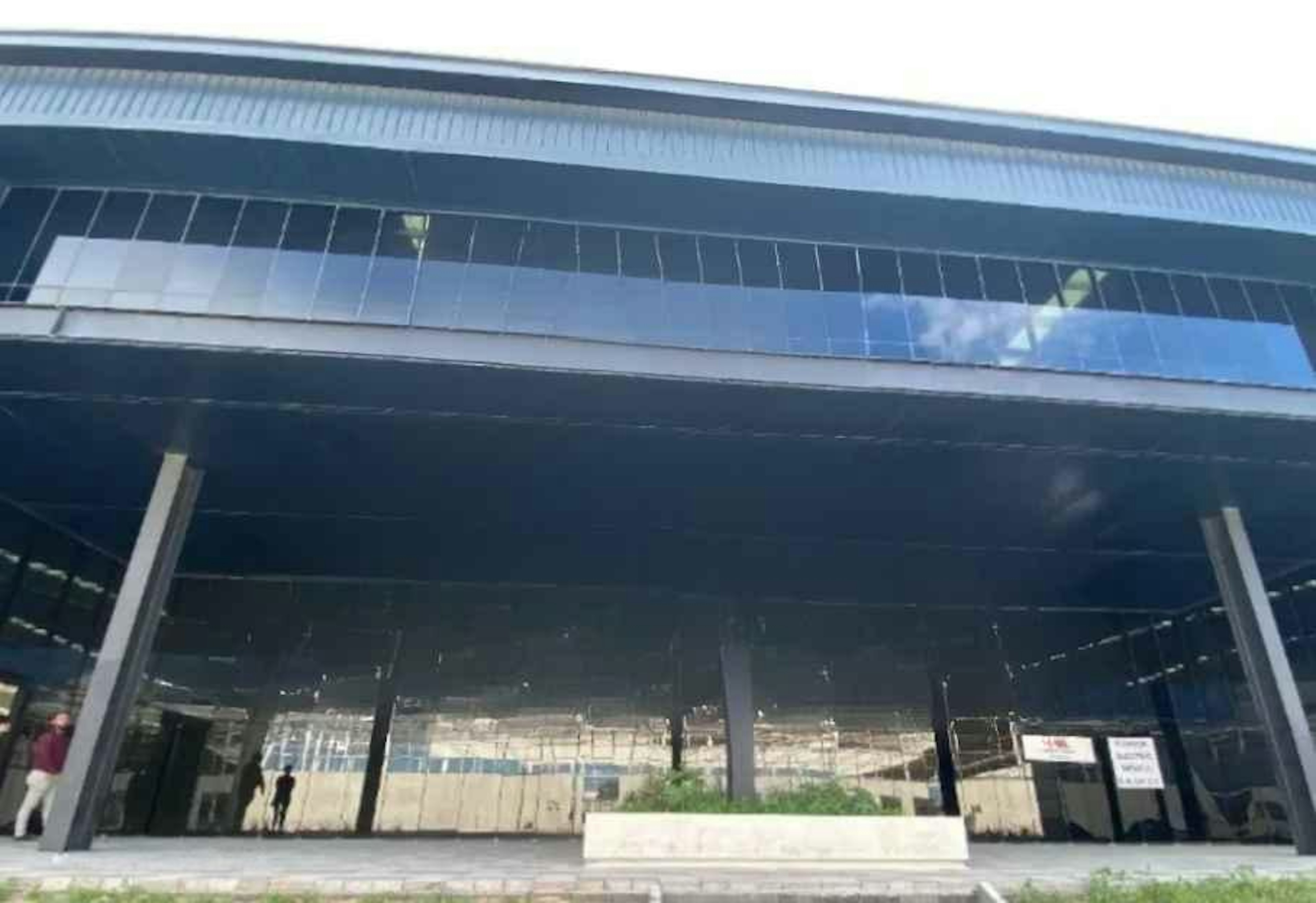 La imagen muestra la fachada de un edificio moderno en construcción o renovación. Se observa una estructura metálica con grandes ventanales y un techo sobresaliente. El edificio parece estar en una etapa avanzada de construcción, con algunas áreas aún sin terminar. La arquitectura sugiere un espacio comercial o de oficinas, con un diseño contemporáneo que enfatiza la transparencia y la luz natural. El entorno indica que podría ser parte de un desarrollo inmobiliario más amplio.