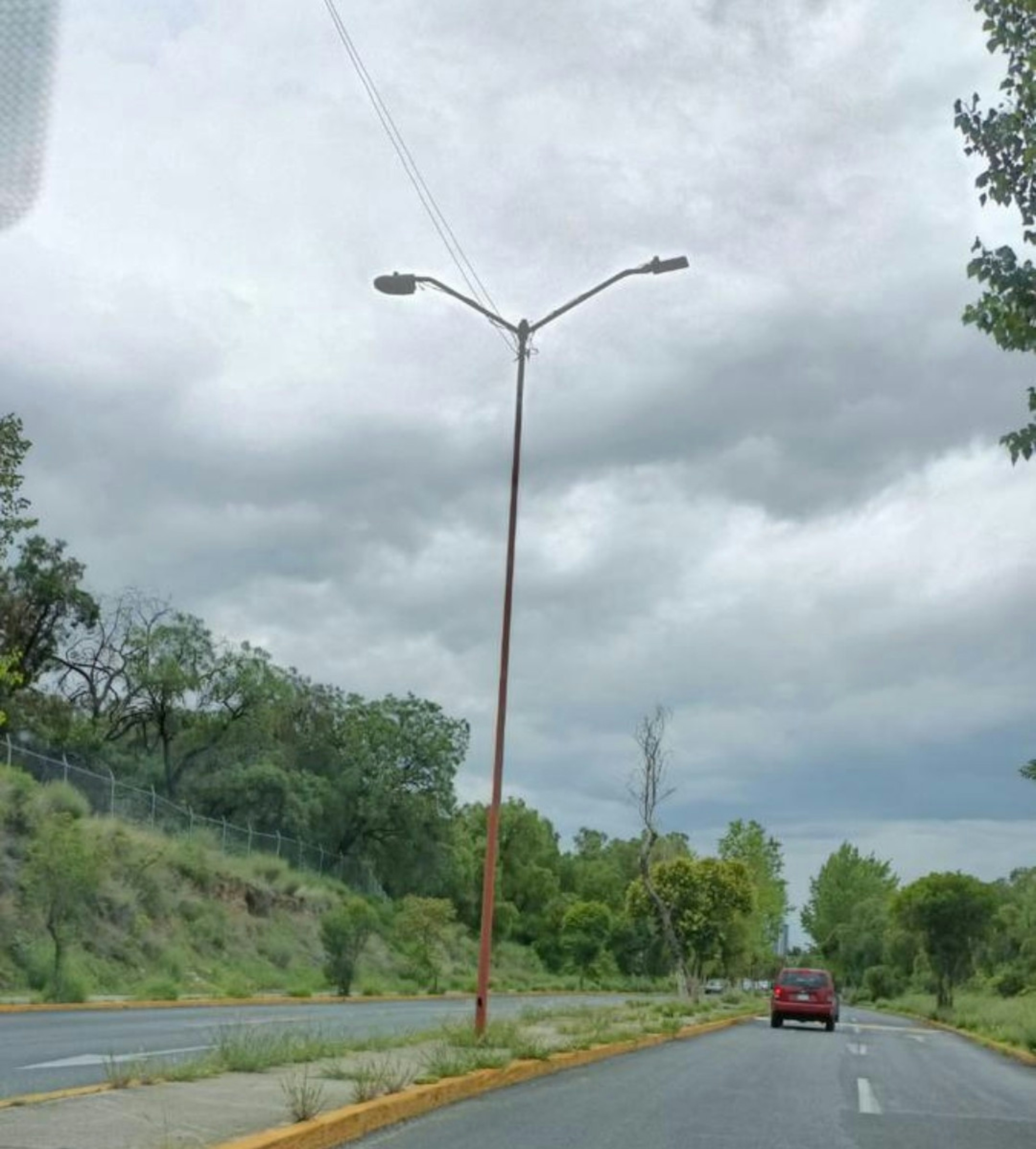 La imagen muestra una carretera en una zona suburbana o rural con vegetación abundante. Un poste de alumbrado público domina el centro de la imagen, mientras que un automóvil rojo se ve a lo lejos. El cielo está nublado y gris, sugiriendo un clima potencialmente lluvioso. La carretera parece estar en buenas condiciones, con líneas divisorias visibles. Este entorno podría ser atractivo para desarrollos residenciales o comerciales de baja densidad.