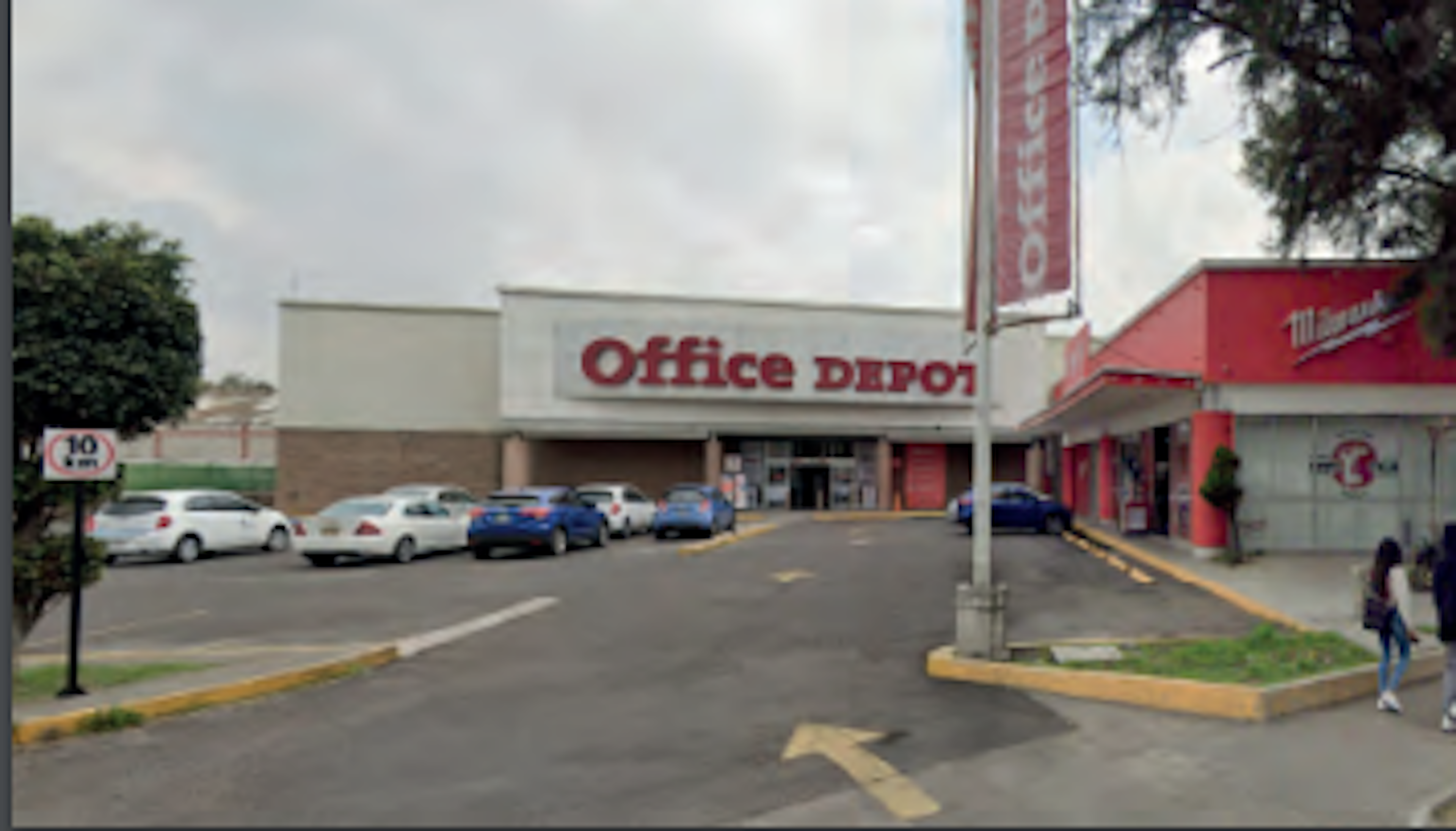 La imagen muestra un local comercial de Office Depot en una zona comercial. El edificio es de una planta, con fachada blanca y roja. Hay un estacionamiento frente al local con varios autos aparcados. Se observa parte de otro local comercial a la derecha, posiblemente una tienda Mattel. La escena es típica de un centro comercial suburbano o plaza comercial, con áreas verdes y señalización visible.