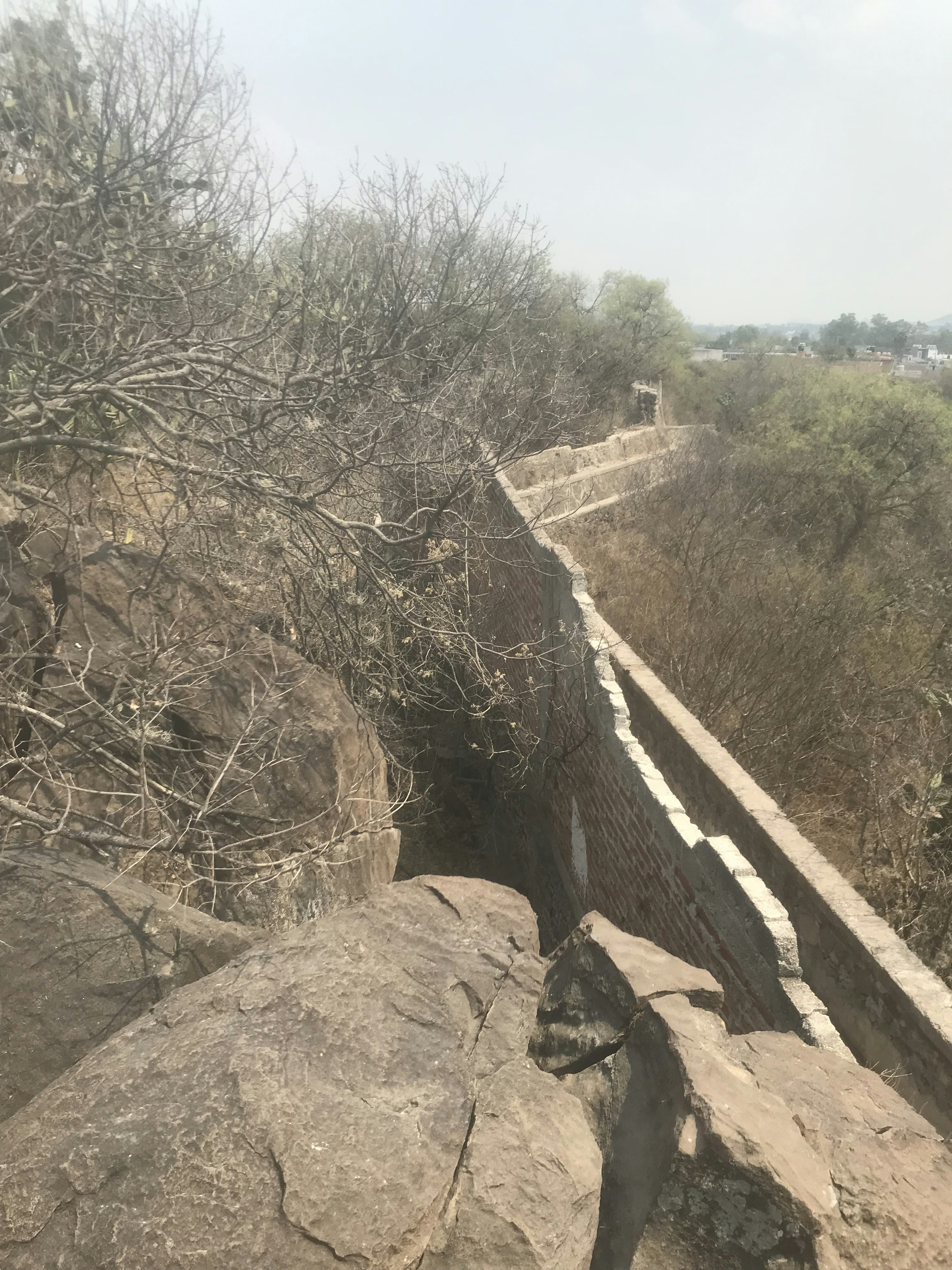 Esta imagen muestra una sección de una antigua muralla o fortificación de piedra, rodeada de vegetación seca y rocas. La estructura se extiende en la distancia, sugiriendo una construcción histórica significativa. El entorno parece árido, con árboles sin hojas y un paisaje desértico. Esta vista podría ser de interés para proyectos de restauración histórica o desarrollo turístico en zonas patrimoniales.