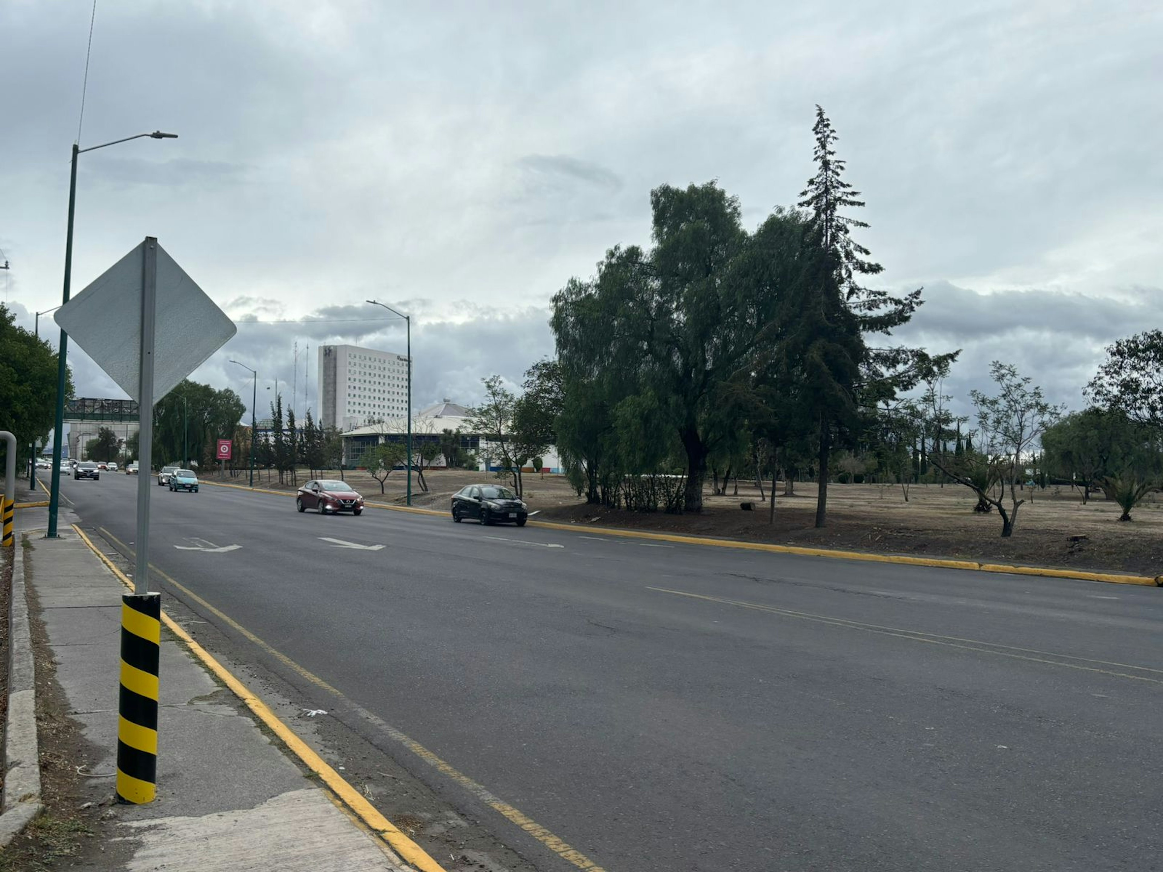 Imagen de una avenida amplia con varios carriles y vehículos en movimiento. Se observa un área verde a un lado, posiblemente un parque o espacio público, y edificaciones en el fondo que podrían ser de interés para desarrollos inmobiliarios.