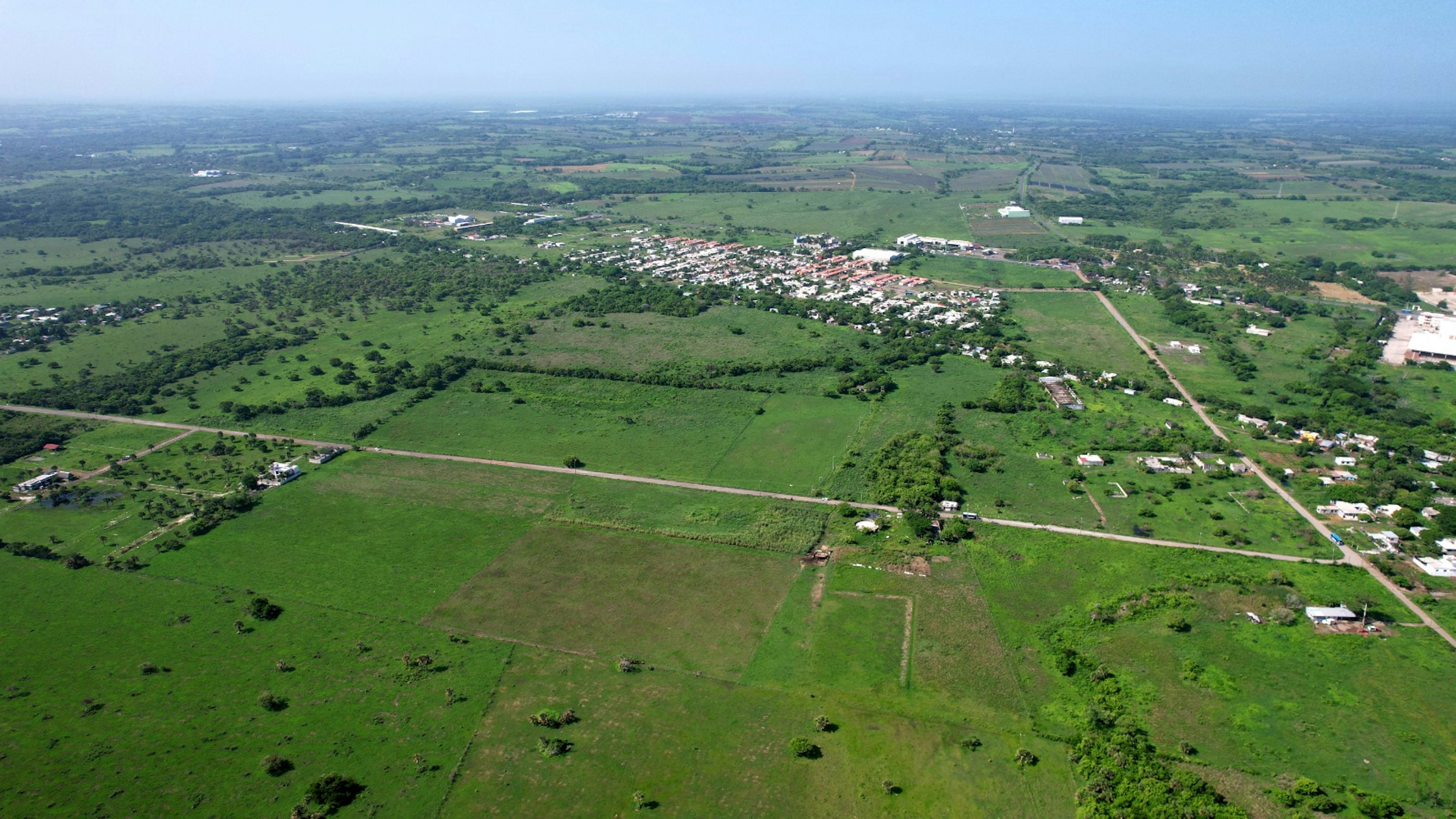 La imagen muestra una vista aérea de una zona rural con extensos campos verdes y una pequeña área urbanizada. Se observan parcelas de terreno, caminos de tierra que las dividen y una concentración de casas con techos rojos en el centro. Esta vista panorámica sugiere potencial para desarrollo inmobiliario, con amplios espacios disponibles para posibles expansiones residenciales o proyectos agrícolas.