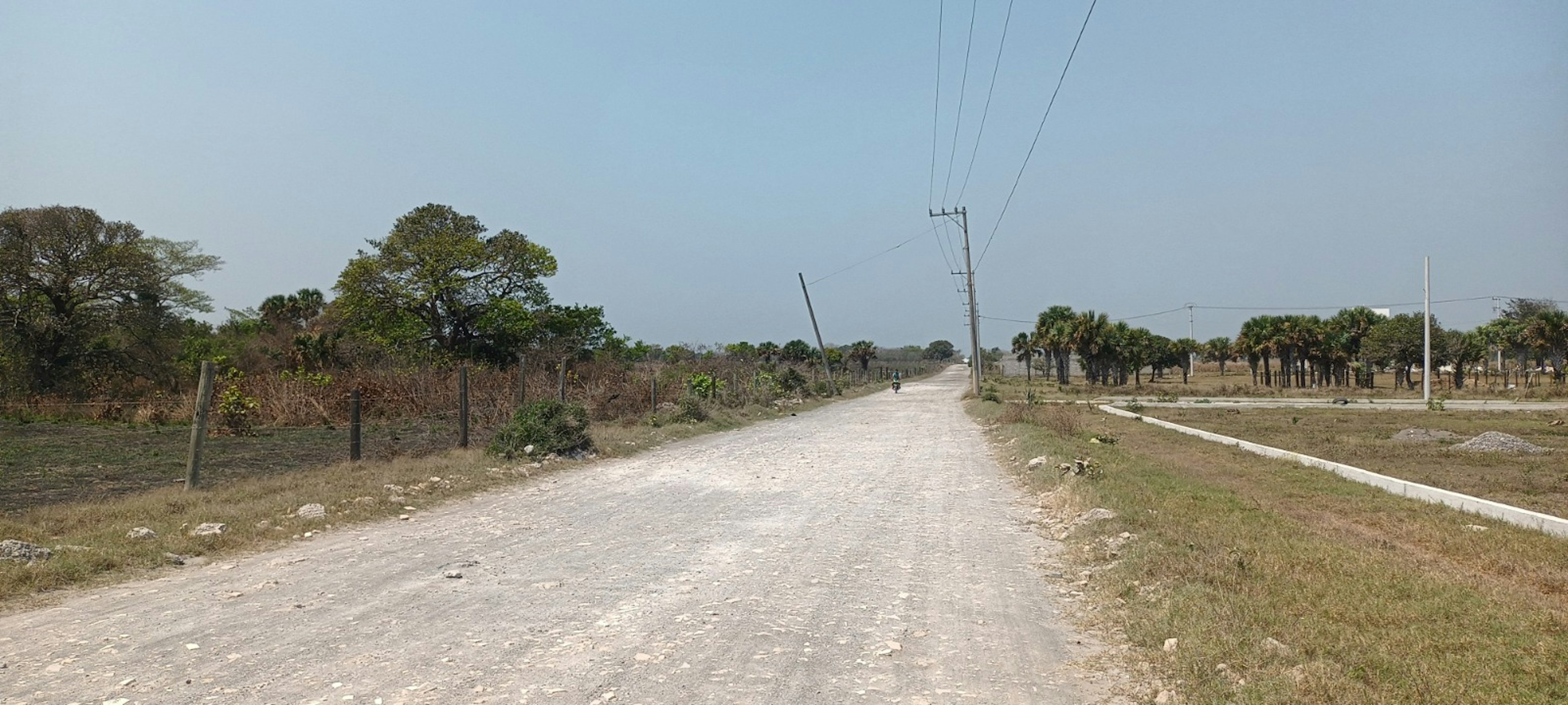 La imagen muestra un camino rural sin pavimentar en un área suburbana en desarrollo. Se observan postes de electricidad a lo largo del camino, vegetación tropical como palmeras y árboles dispersos. El terreno parece estar dividido en lotes, sugiriendo un potencial desarrollo inmobiliario. El cielo es claro y el ambiente general es de una zona en transición entre lo rural y lo urbano, ideal para futuros proyectos residenciales.