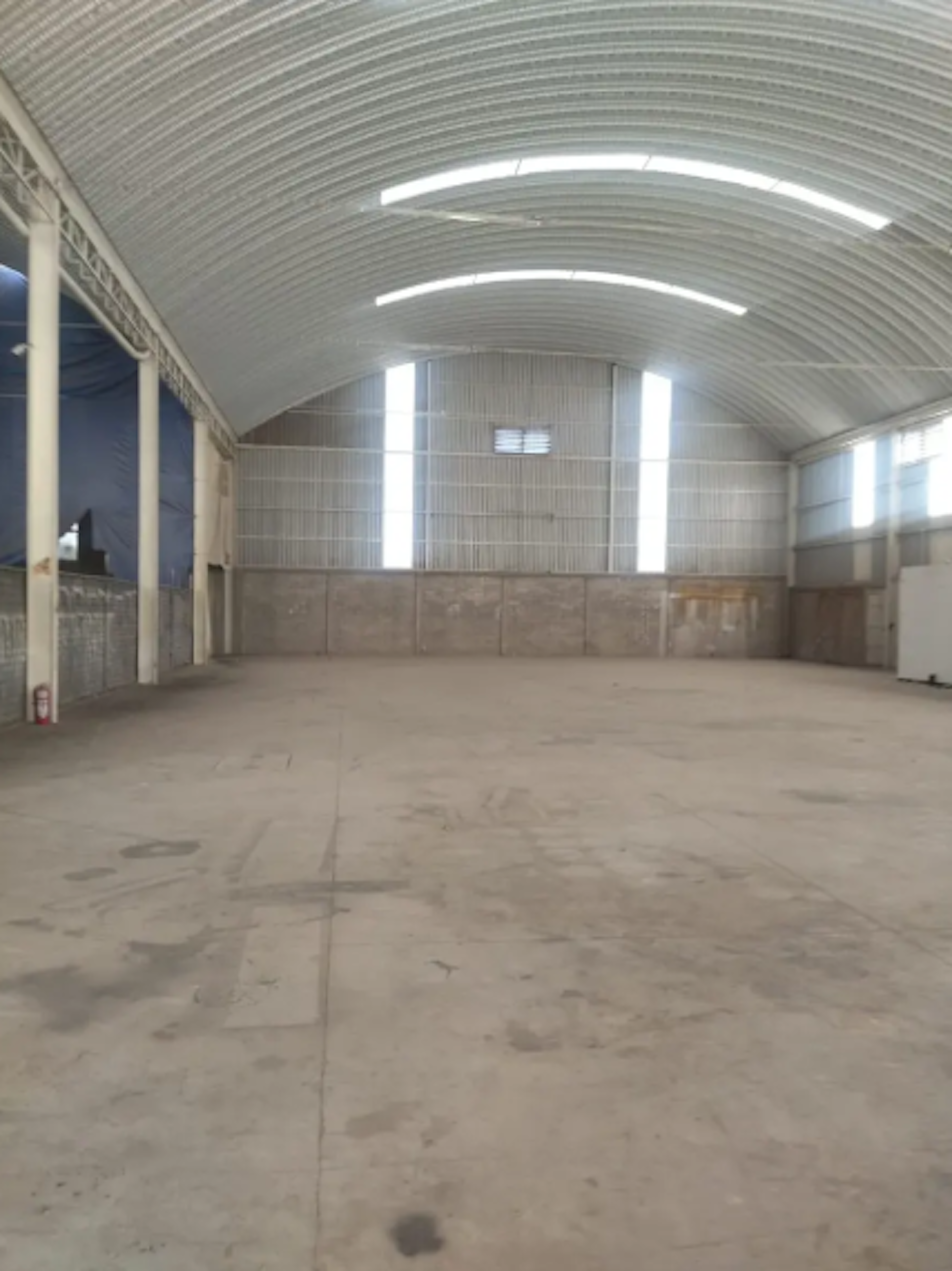La imagen muestra un amplio espacio interior industrial o de almacenamiento. Tiene un techo abovedado alto con iluminación fluorescente. Las paredes son una combinación de concreto en la parte inferior y paneles blancos en la superior. El piso es de concreto sin acabados. El espacio está vacío, lo que sugiere que está disponible para alquiler o remodelación. Es ideal para uso comercial o industrial debido a su gran tamaño y estructura.
