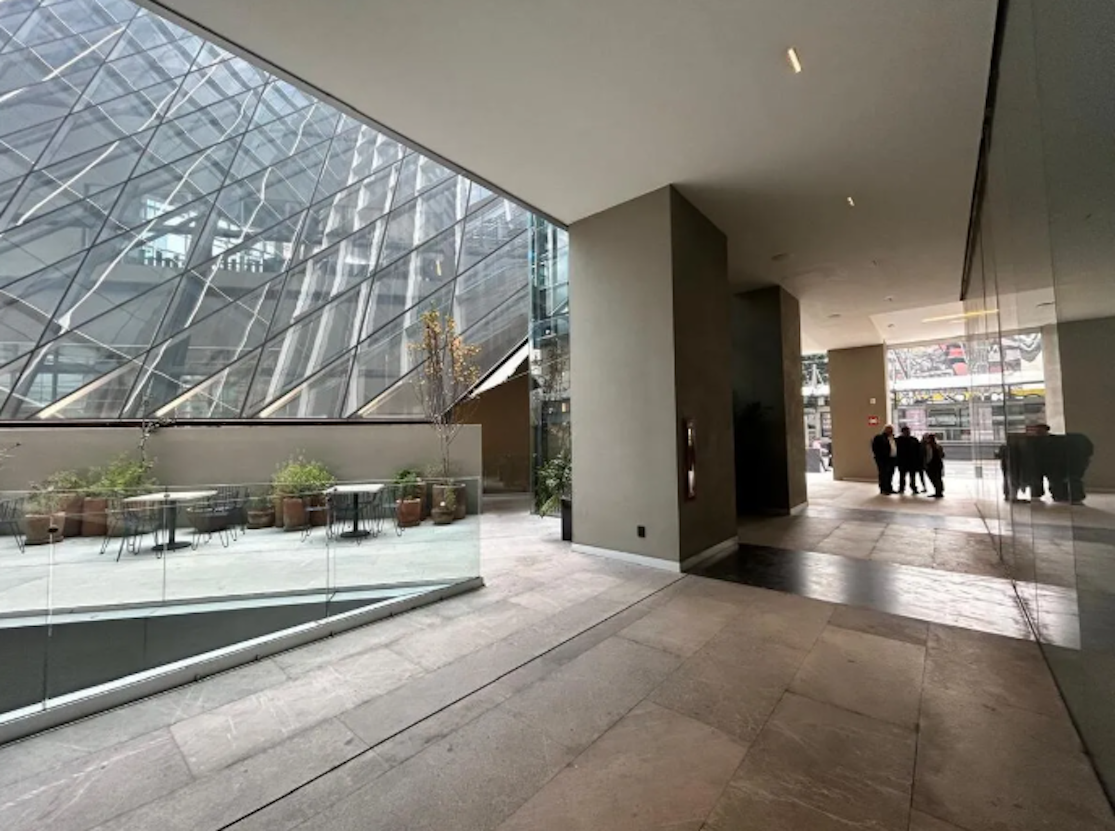 Esta imagen muestra un moderno vestíbulo de un edificio corporativo o comercial. Presenta una impresionante estructura de vidrio con diseño geométrico que permite la entrada de luz natural. El espacio interior es amplio, con pisos de mármol, columnas elegantes y áreas de descanso con plantas. Se observa una combinación de diseño arquitectónico contemporáneo y funcionalidad, típico de propiedades inmobiliarias de alto nivel.