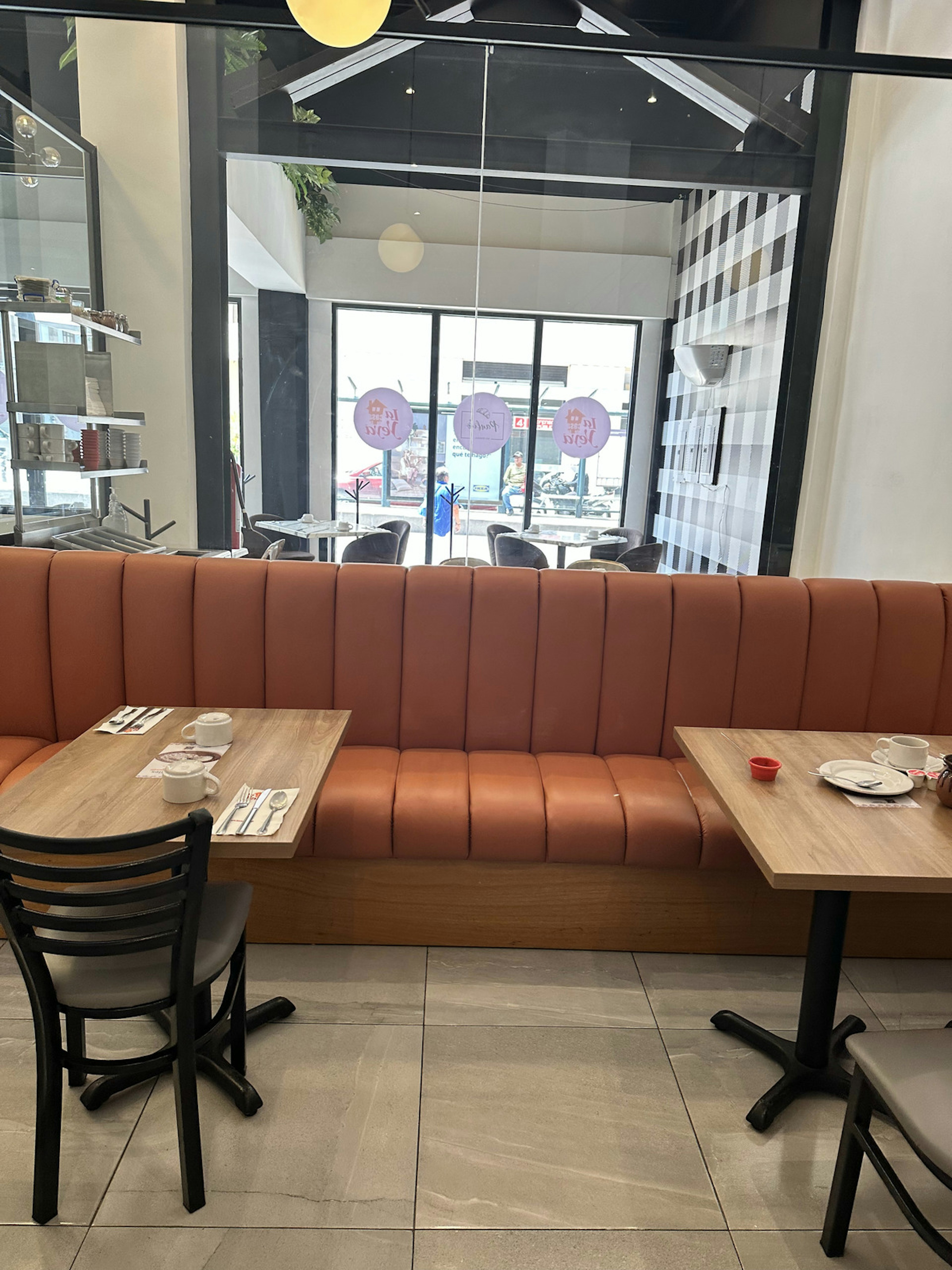 Interior de un local comercial, posiblemente una cafetería o restaurante, con una banqueta larga de color terracota, mesas de madera, sillas negras y decoración moderna. Grandes ventanas permiten la entrada de luz natural.