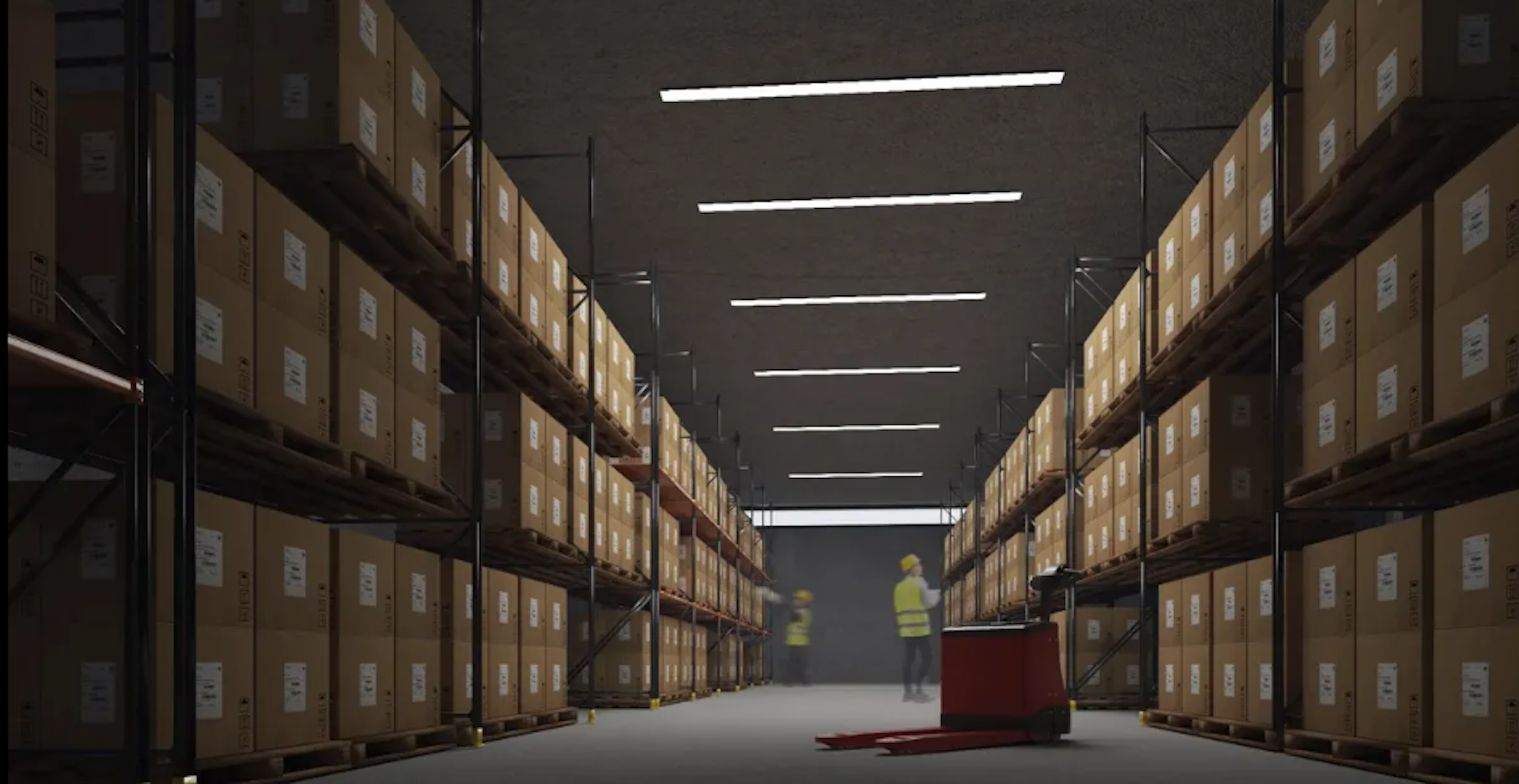 La imagen muestra un amplio almacén o centro de distribución con altos estantes metálicos llenos de cajas de cartón etiquetadas. Las luces fluorescentes iluminan el largo pasillo central. Al fondo se ven dos figuras con chalecos de seguridad amarillos. En primer plano hay un palet rojo. El espacio refleja una operación logística eficiente y organizada, típica de grandes empresas de comercio electrónico o cadenas de suministro.