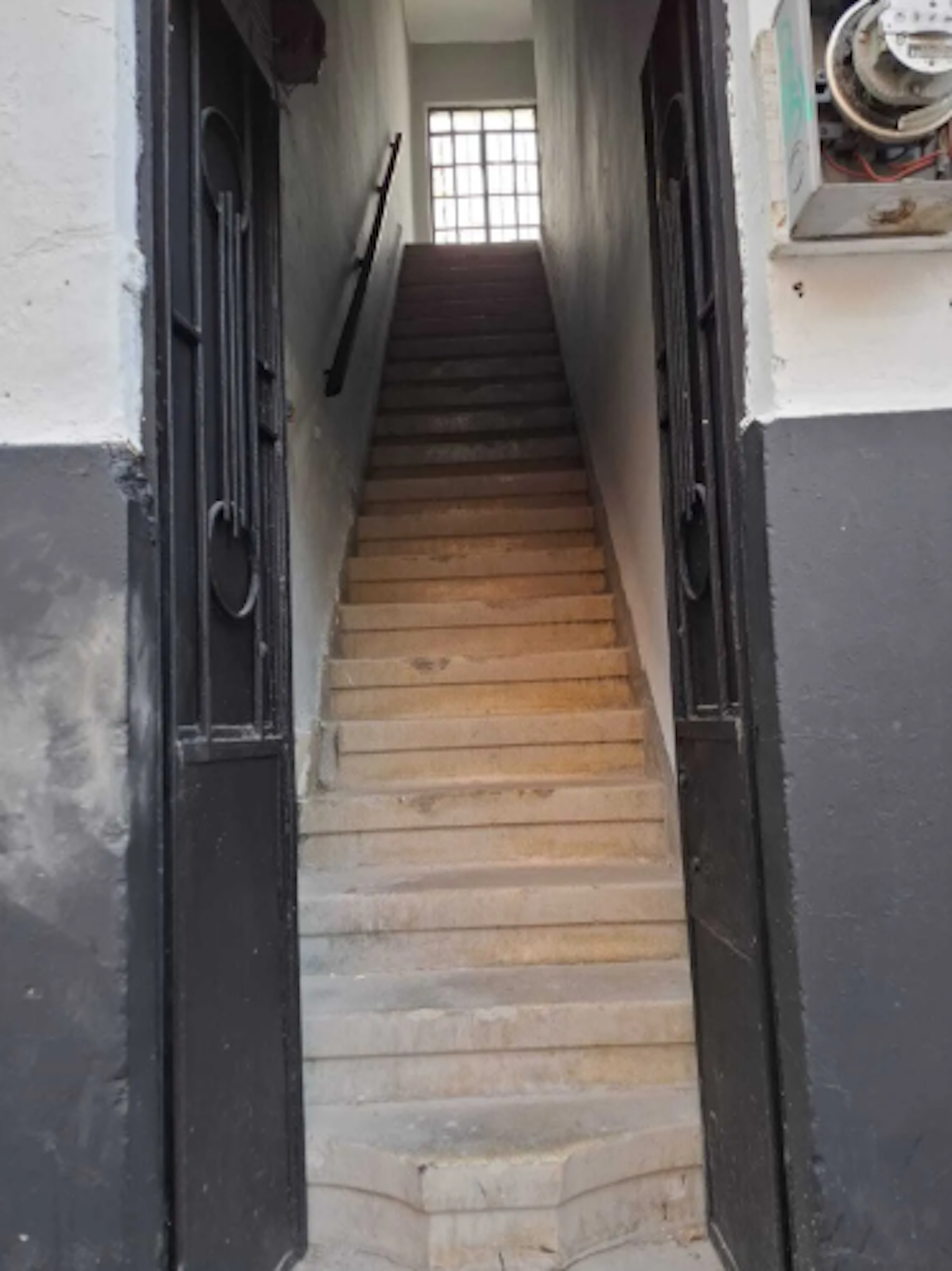 La imagen muestra una entrada estrecha con escaleras de madera que conducen al segundo piso de un edificio antiguo. Las paredes son blancas y hay una barandilla negra. La puerta de entrada es negra y ornamentada. Al final de las escaleras se ve una ventana que permite la entrada de luz natural. El espacio parece necesitar algunas reparaciones y mantenimiento.