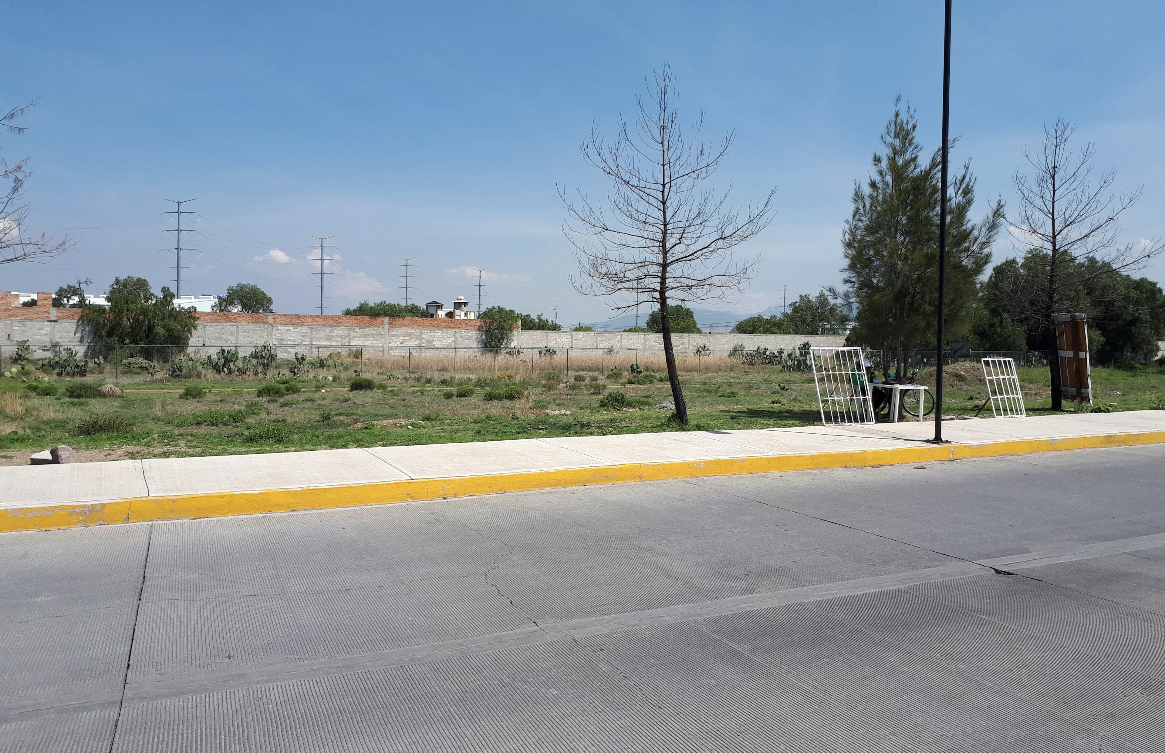 Terreno despejado con vegetación baja y algunos árboles, ubicado en una zona urbana con infraestructura básica como aceras y alumbrado público. Adecuado para desarrollo inmobiliario.