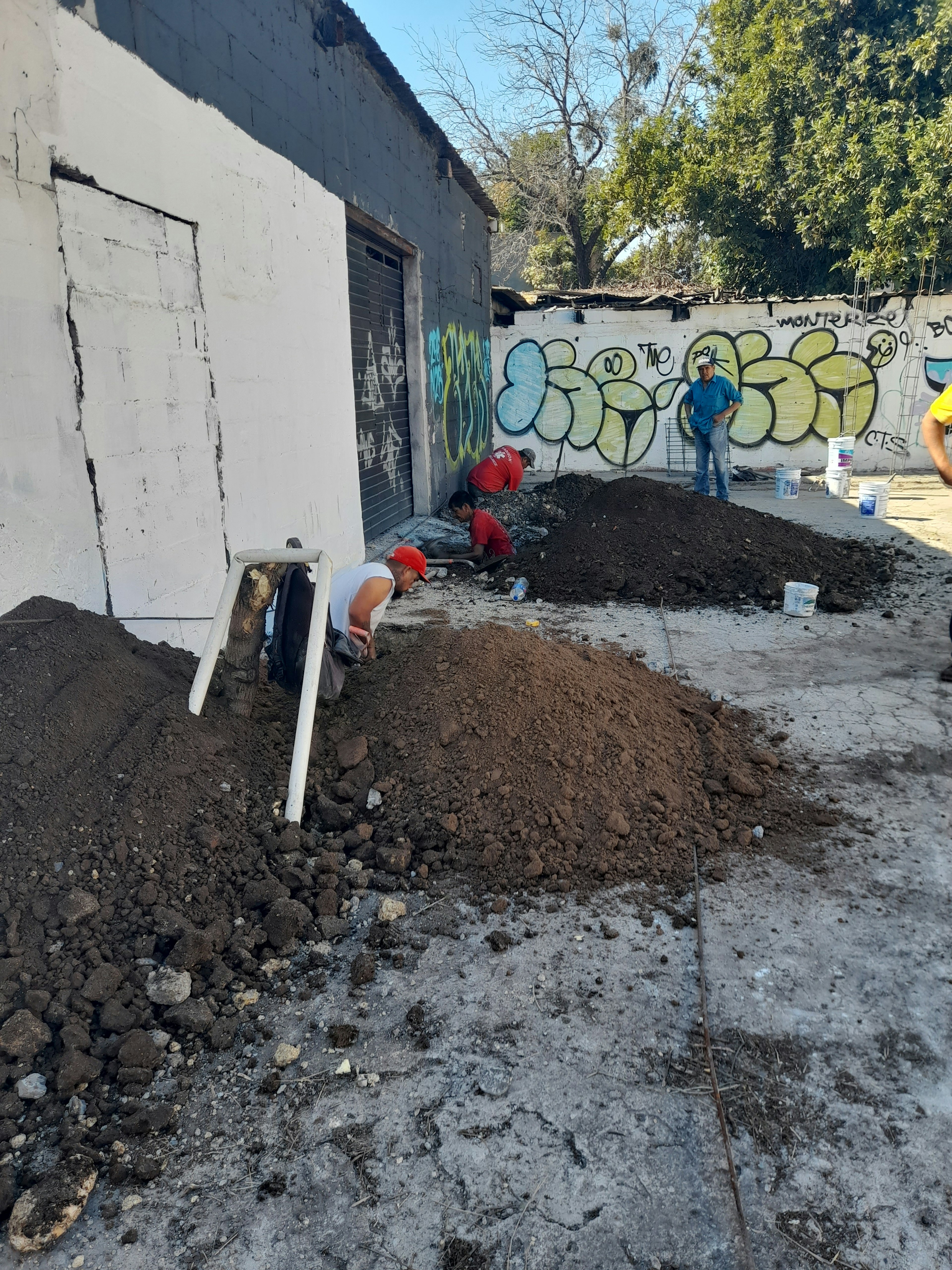 La imagen muestra una escena de construcción o renovación en un área urbana. Se observa una pared blanca con grafitis y varios montículos de tierra y escombros. Hay personas trabajando en el sitio, posiblemente en tareas de excavación o preparación del terreno. El entorno sugiere una zona en proceso de remodelación o mejora, posiblemente para un proyecto inmobiliario o de renovación urbana.