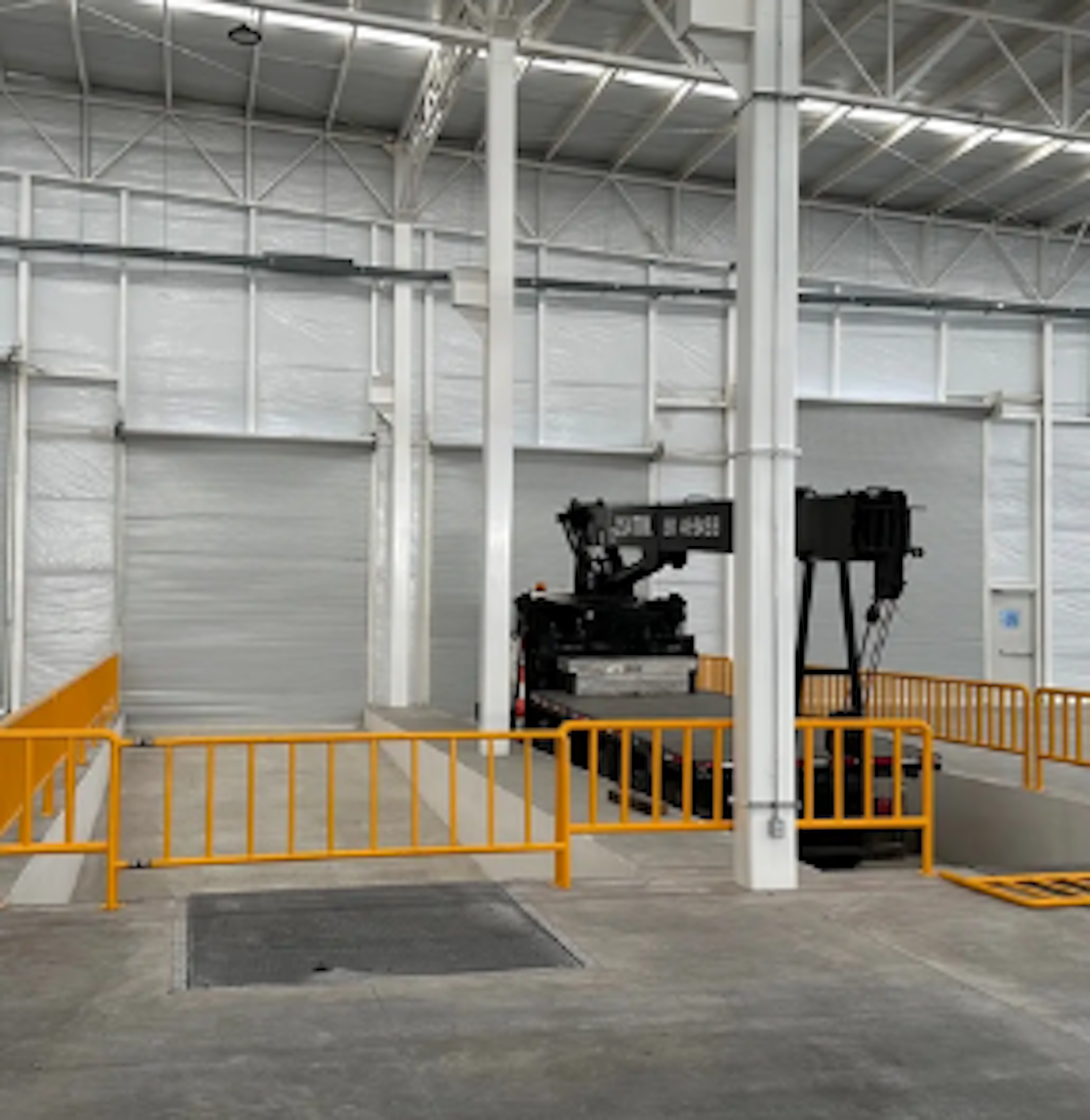 Interior de un almacén industrial con techos altos y estructura metálica. Se observa una montacargas estacionada junto a barandillas de seguridad amarillas. Iluminación artificial y puertas de enrollar metálicas.