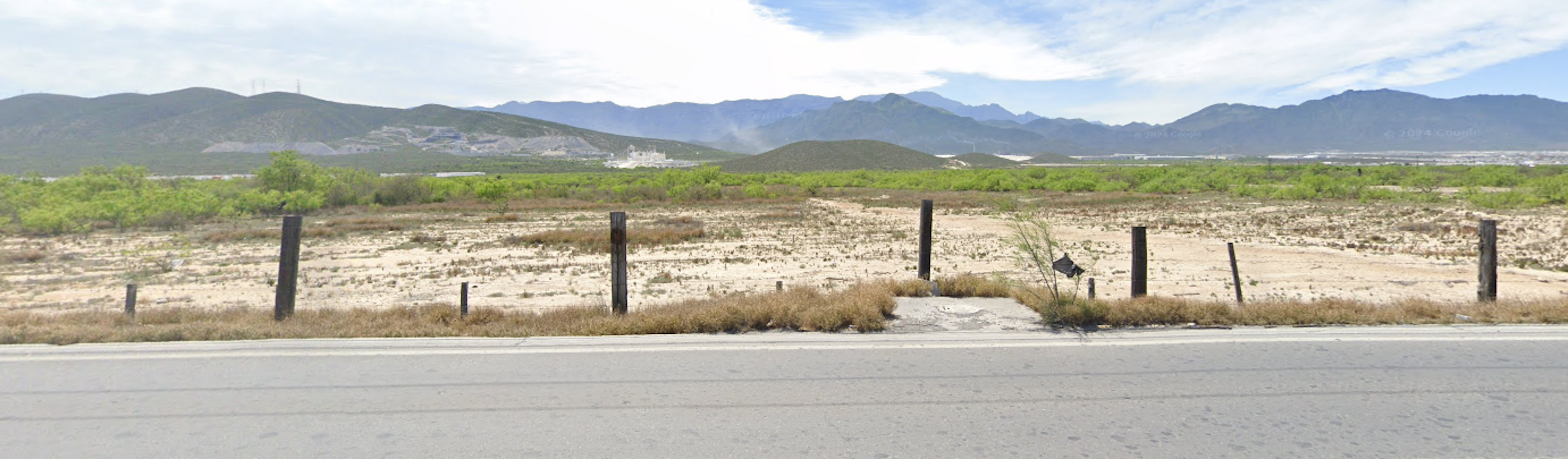 La imagen muestra un terreno árido y sin desarrollar en un área rural o suburbana. Se ve una carretera en primer plano, seguida de una cerca y un vasto terreno desértico con vegetación escasa. Al fondo se aprecian montañas y un cielo parcialmente nublado. El terreno parece tener potencial para desarrollo inmobiliario, aunque actualmente está sin construir y en estado natural.