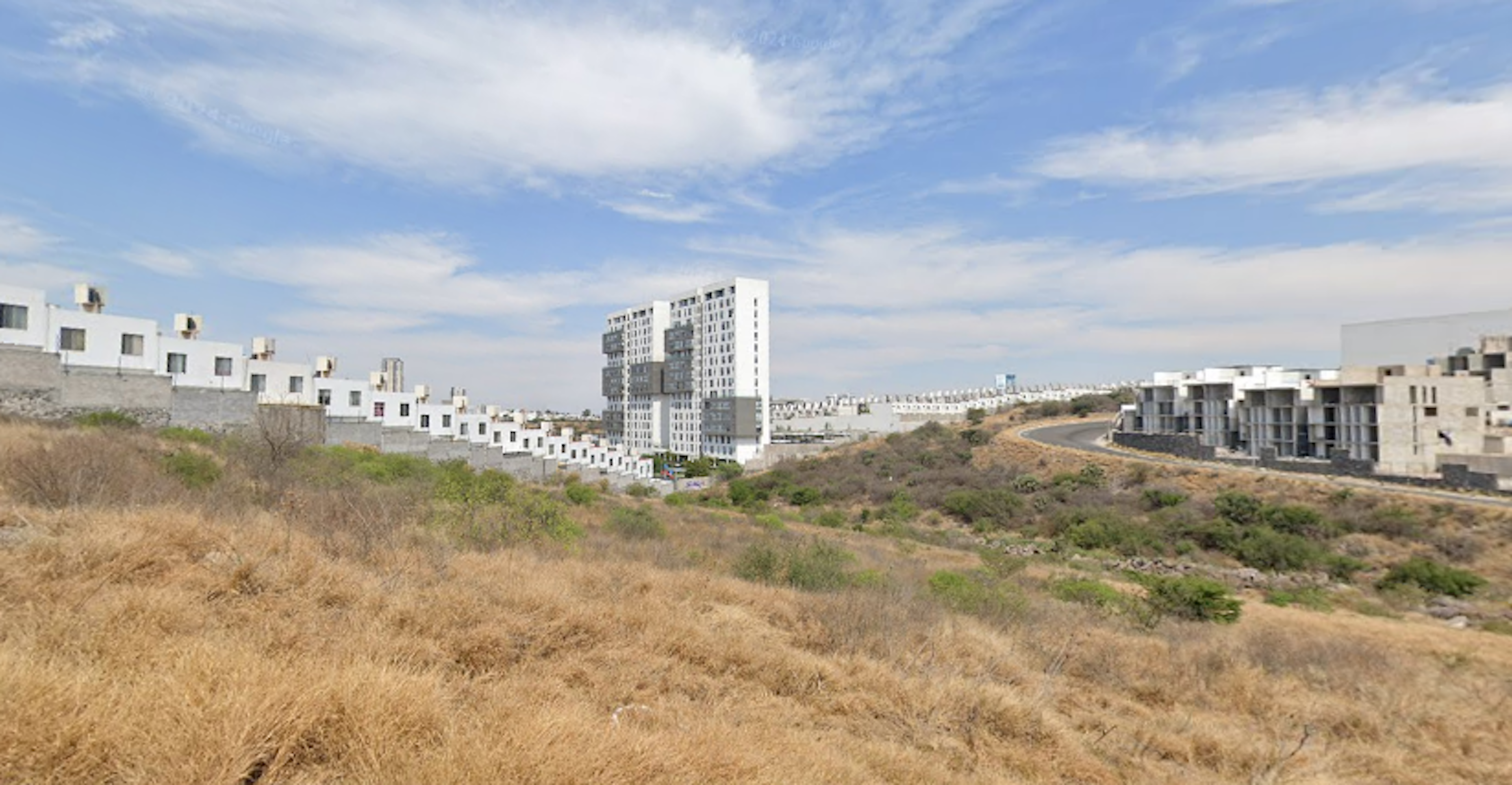 La imagen muestra un desarrollo urbano en construcción, con edificios de apartamentos blancos y altos a la izquierda y derecha. En el centro se destaca un complejo de torres residenciales más grandes. El terreno en primer plano es árido, con vegetación seca. El cielo es azul con algunas nubes, sugiriendo un clima seco. La escena refleja una expansión inmobiliaria en una zona anteriormente no desarrollada.