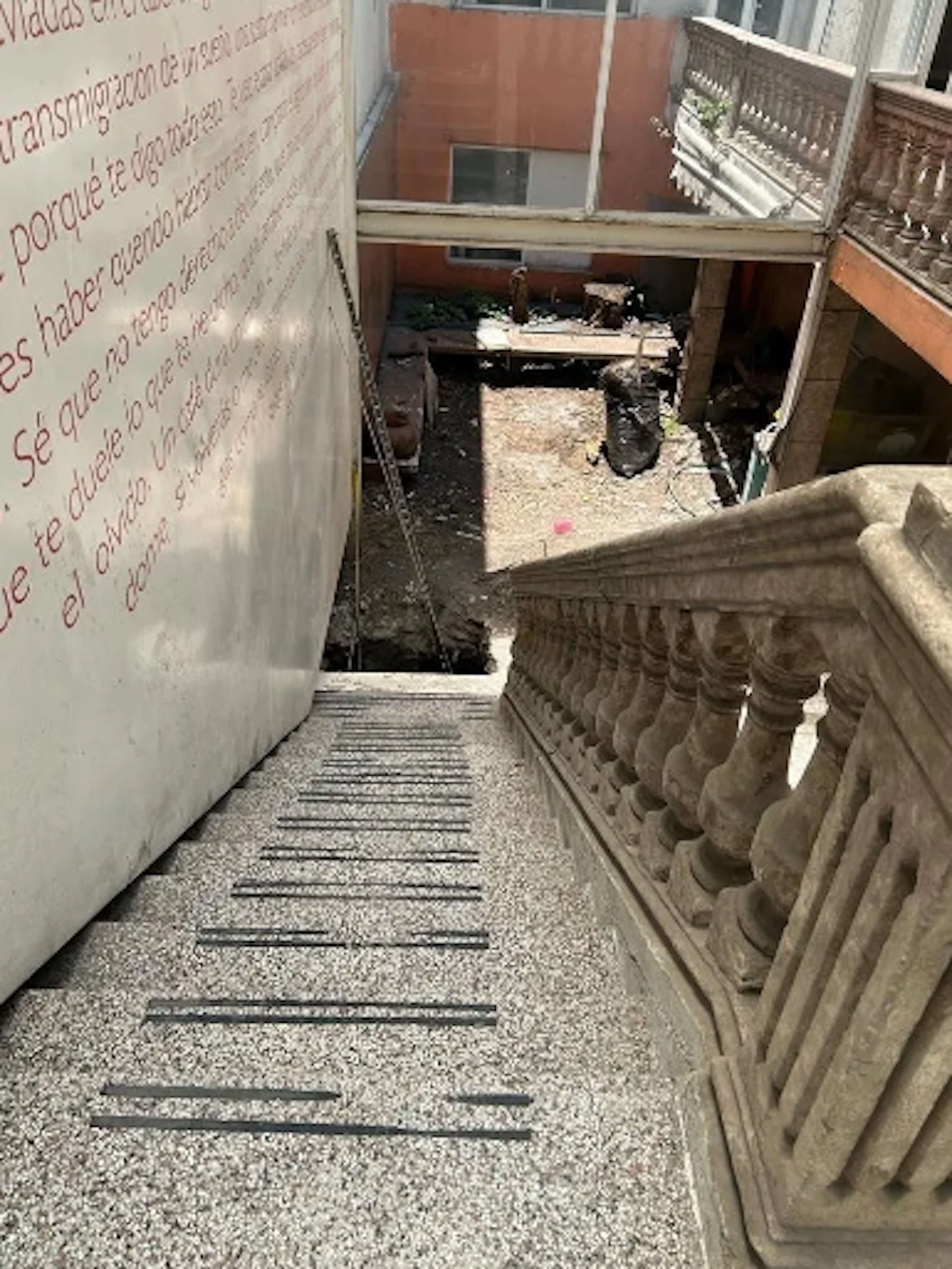 La imagen muestra un área en construcción o remodelación de un edificio. Se ve una escalera improvisada con barras metálicas sobre un suelo de granito. A la izquierda hay una pared con texto en español, y a la derecha una barandilla ornamental. Al fondo se aprecia un patio interior con escombros y materiales de construcción. El espacio parece estar en proceso de renovación.