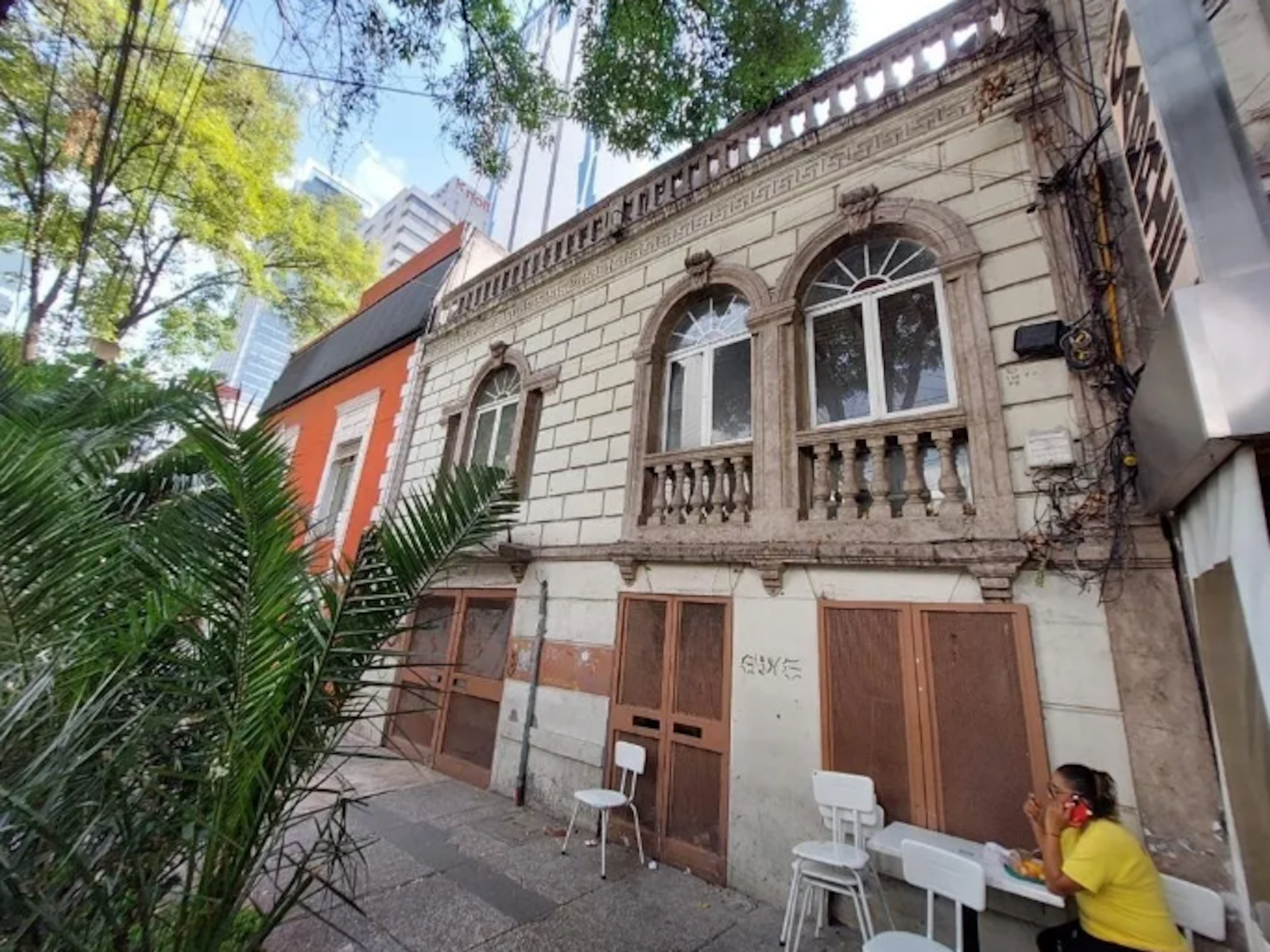 La imagen muestra un edificio antiguo de estilo neoclásico con detalles ornamentales, balcones y ventanas arqueadas. El primer piso parece estar cerrado con puertas de madera. Junto a él hay un edificio naranja más moderno. Se ven palmeras y árboles en primer plano, y edificios altos al fondo, sugiriendo una zona urbana en transición. Hay sillas blancas en la acera, indicando posible uso comercial.