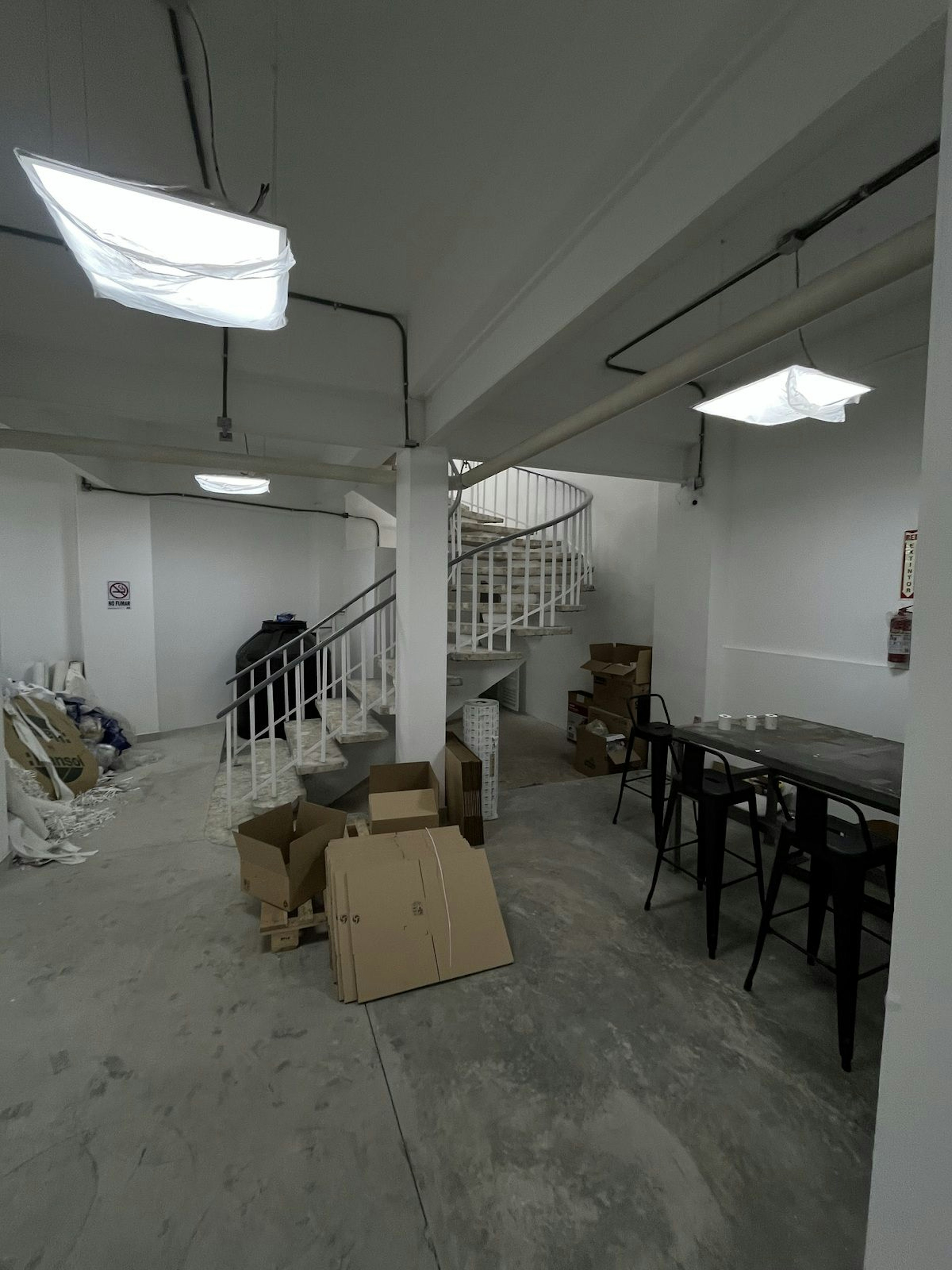La imagen muestra un espacio interior industrial en proceso de renovación. Se observa una escalera de caracol blanca, paredes sin acabados, suelo de cemento y cajas de cartón dispersas. Hay luces fluorescentes colgantes con difusores de tela. Se ven algunas mesas altas negras y un extintor en la pared. El espacio parece estar en transición, posiblemente convirtiéndose en un área de oficinas o un espacio comercial.
