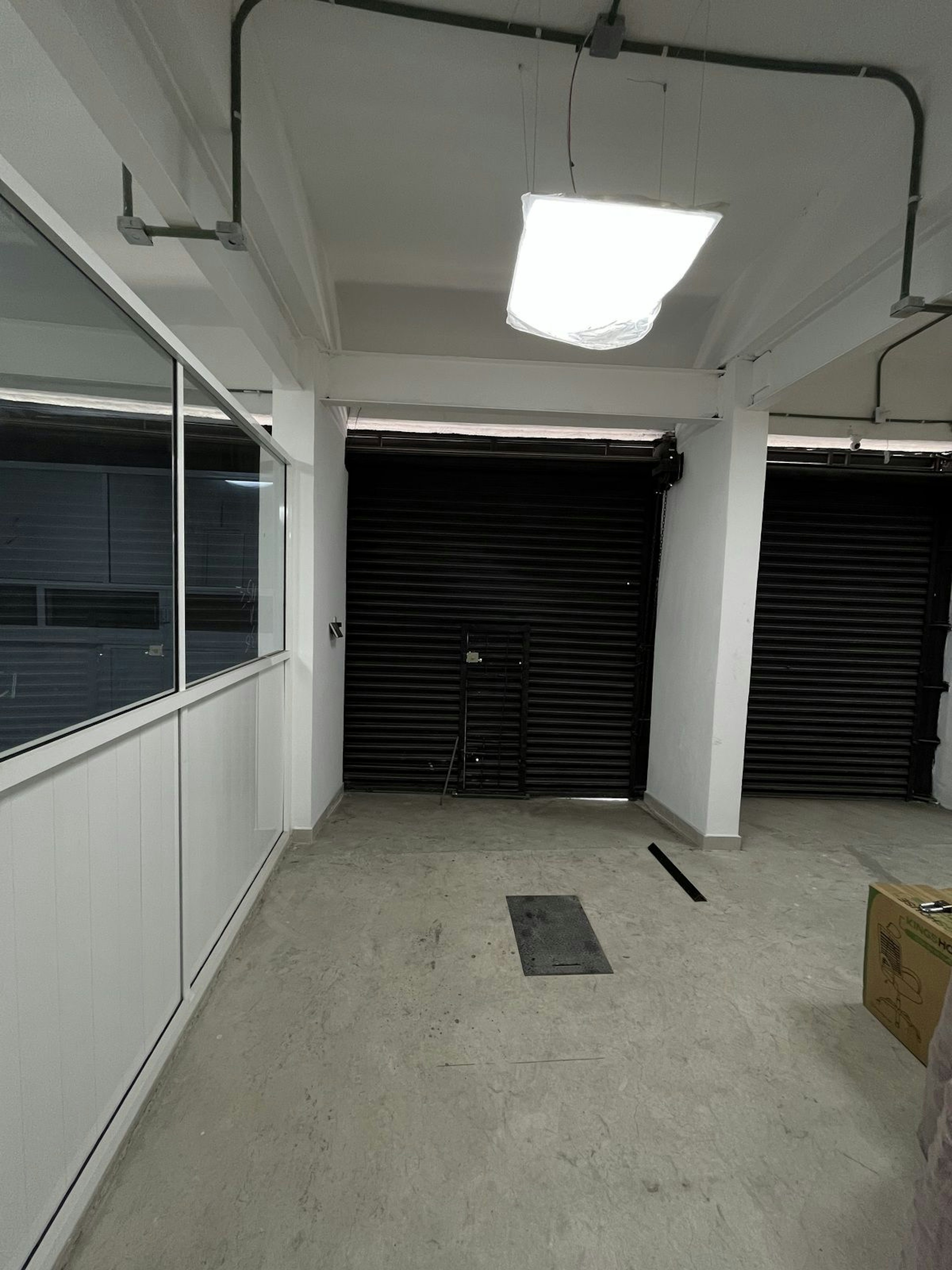 La imagen muestra un espacio interior industrial o comercial en construcción o renovación. Tiene paredes blancas, un piso de concreto sin terminar, y una ventana grande a la izquierda. Al fondo hay puertas enrollables negras. El techo tiene tuberías y una luz LED cuadrada colgando. El espacio parece estar en proceso de ser acondicionado, posiblemente para una oficina o almacén.