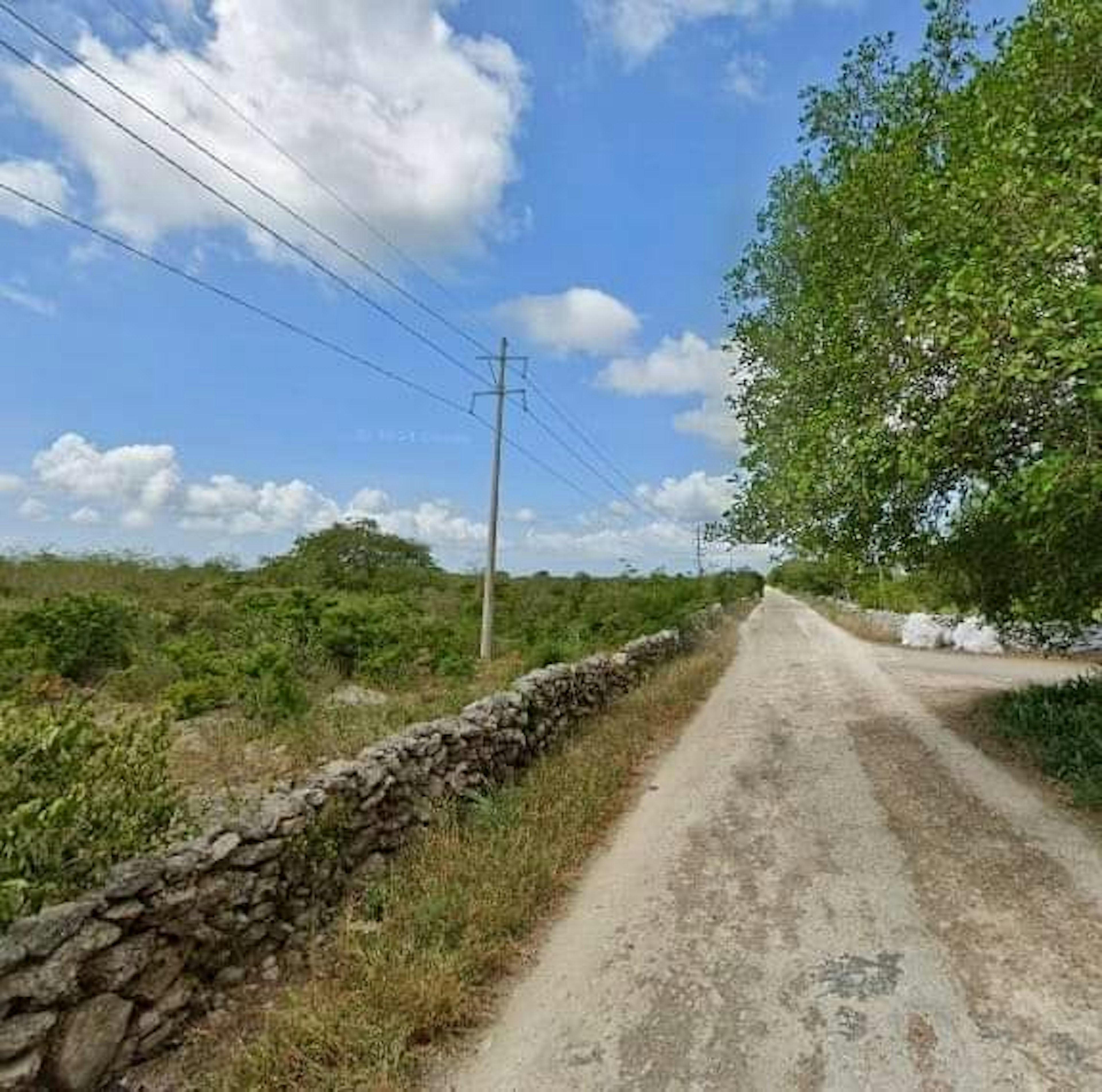 La imagen muestra un camino rural de tierra bordeado por muros de piedra bajos. A un lado se observa vegetación exuberante y árboles. El cielo está despejado con algunas nubes blancas. Se ven postes eléctricos a lo largo del camino, sugiriendo que la zona tiene acceso a electricidad. El paisaje parece tranquilo y poco desarrollado, potencialmente atractivo para propiedades rurales o fincas.