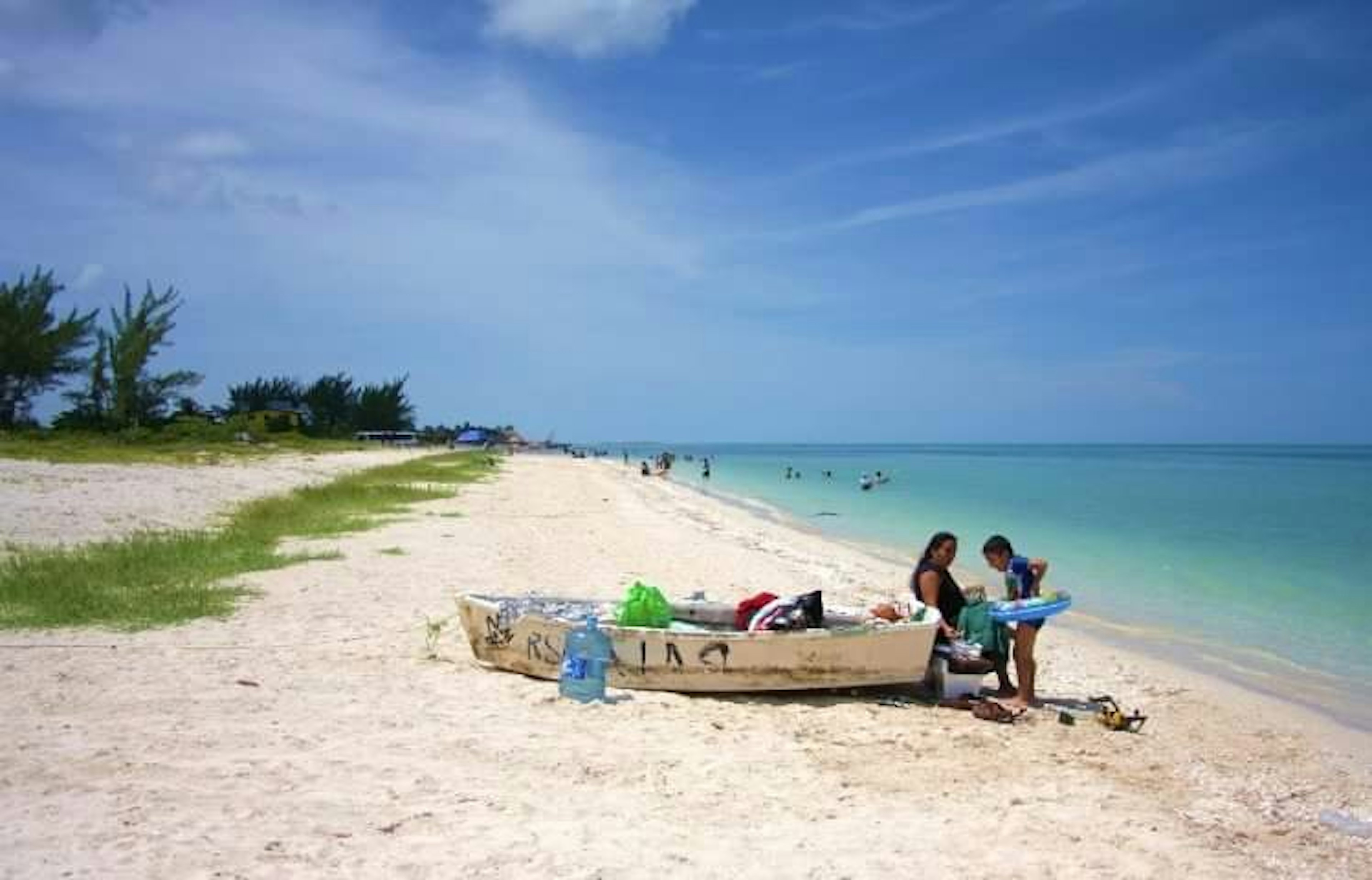 Esta imagen muestra una hermosa playa tropical con arena blanca y aguas cristalinas de color turquesa. Se ve un bote de madera en la orilla, junto a algunos objetos de playa. La playa se extiende a lo largo, con vegetación y algunas estructuras visibles en la distancia. Hay personas disfrutando del mar y la playa. El cielo es azul con algunas nubes, creando un ambiente paradisíaco perfecto para propiedades frente al mar.