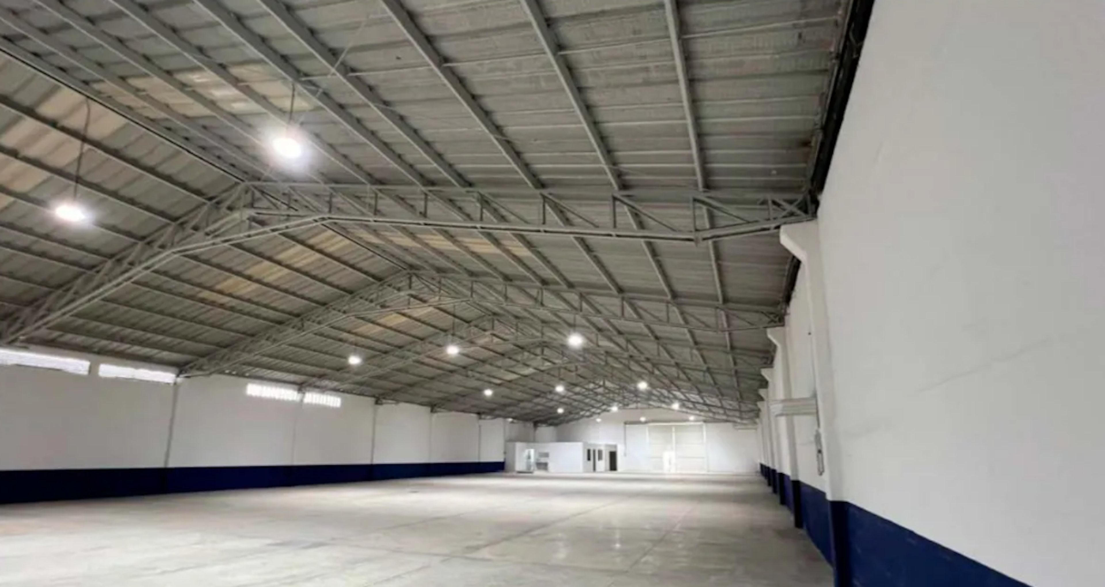 La imagen muestra un amplio espacio industrial o de almacenamiento. Se observa un techo alto con estructura metálica y luces colgantes. Las paredes son blancas con una franja azul en la parte inferior. El piso es de concreto pulido, reflejando la iluminación. Al fondo se ven puertas y ventanas, sugiriendo áreas adicionales. El espacio está vacío, limpio y bien iluminado, ideal para uso comercial o industrial.