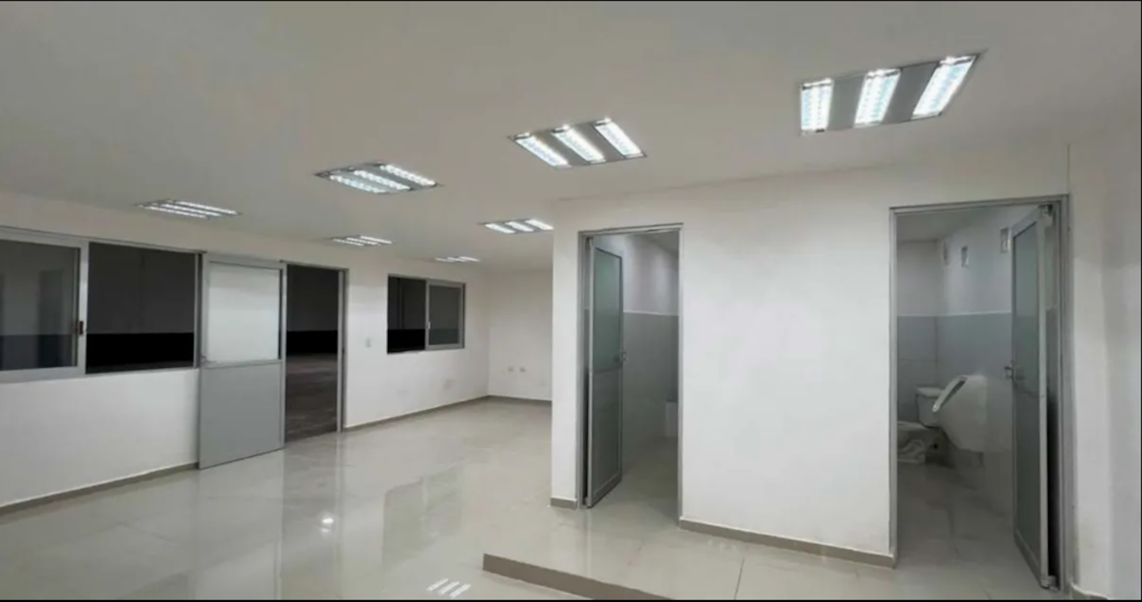 La imagen muestra un espacio de oficina moderno y luminoso. Tiene paredes blancas, piso brillante y varios paneles de iluminación en el techo. Hay múltiples puertas corredizas de vidrio, algunas con persianas. Se ven dos entradas más grandes a la derecha, posiblemente salas de reuniones o áreas privadas. El diseño es minimalista y funcional, ideal para un entorno de trabajo profesional.