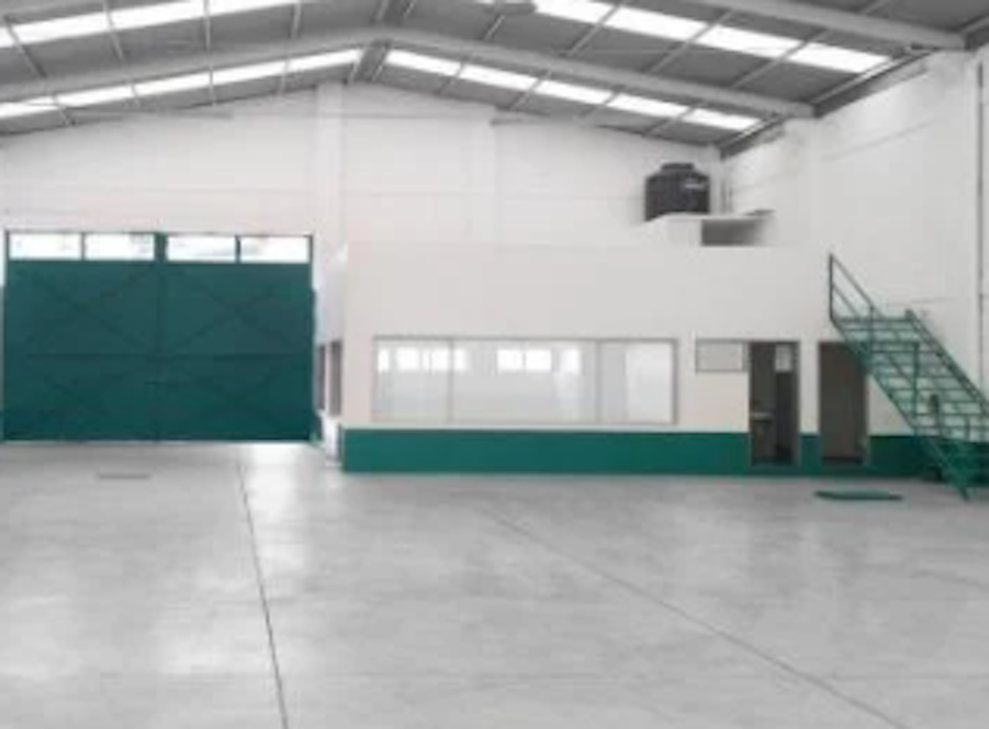 La imagen muestra un amplio espacio interior industrial o comercial. Tiene techos altos con iluminación fluorescente, paredes blancas con detalles en verde, y un piso de concreto. Se observa una gran puerta verde, posiblemente de tipo enrollable, y una escalera metálica verde en un lateral. El espacio parece ser una bodega o área de almacenamiento vacía, lista para ser utilizada o adaptada para diversos fines comerciales o industriales.