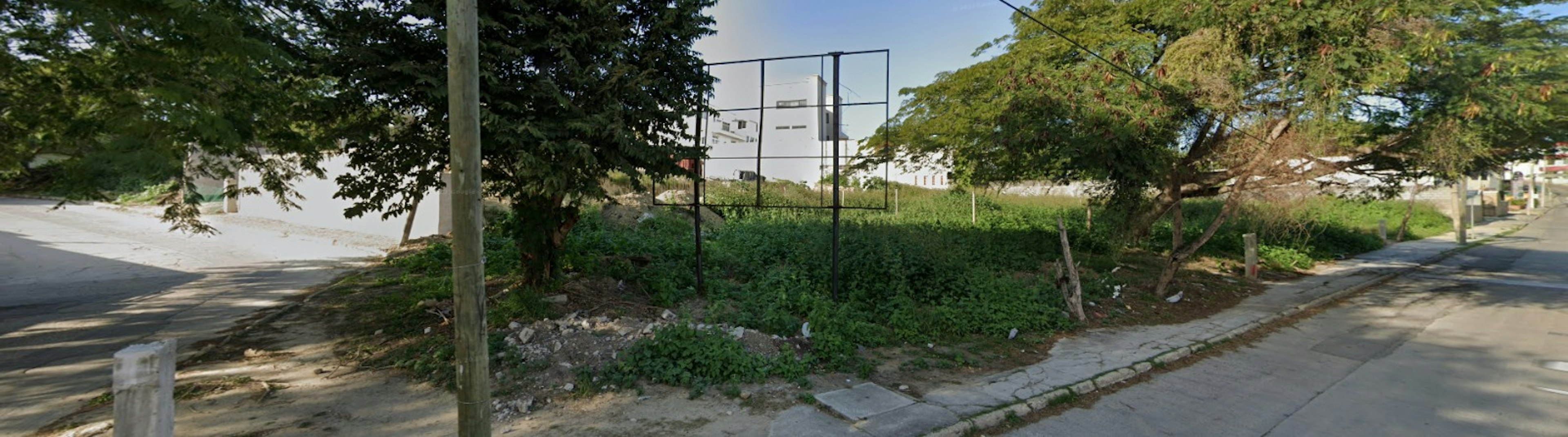 La imagen muestra un terreno urbano sin desarrollar en una esquina. Está cubierto de vegetación silvestre y tiene una estructura metálica, posiblemente un tablero de baloncesto o una valla. El terreno está rodeado por aceras y calles pavimentadas, con algunos árboles maduros proporcionando sombra. Hay edificios visibles en el fondo, sugiriendo que se encuentra en un área residencial o mixta. El lote parece desatendido, con algunos escombros visibles, pero tiene potencial para desarrollo inmobiliario.